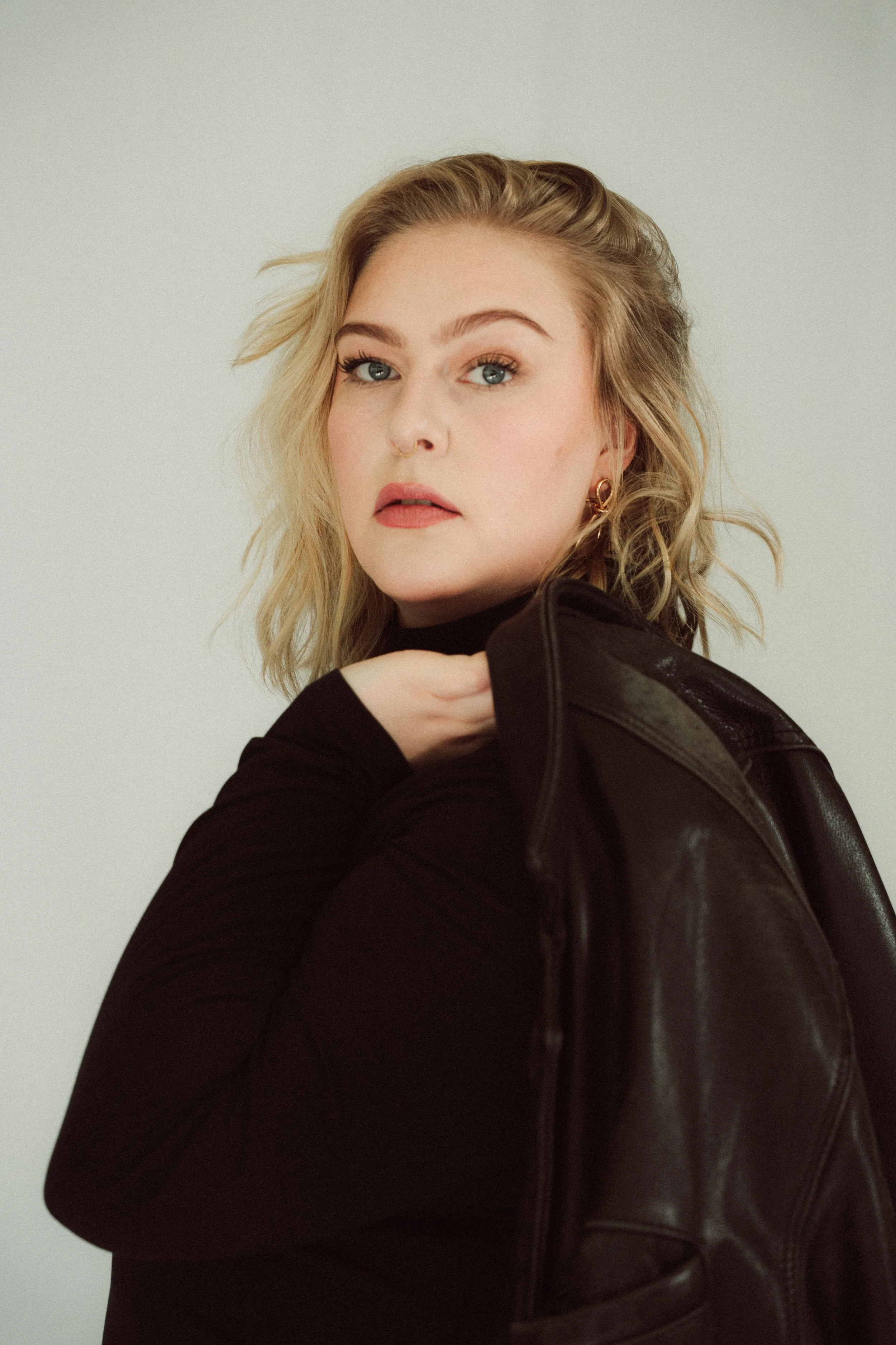 Portrait einer jungen Frau mit blonden, mittellangen, gelockten Haaren, blauen Augen und rot-lippen, trägt eine schwarze Spitze, Lederjacke, und goldene Ohrringe, schaut nach links, vor neutralem Hintergrund.