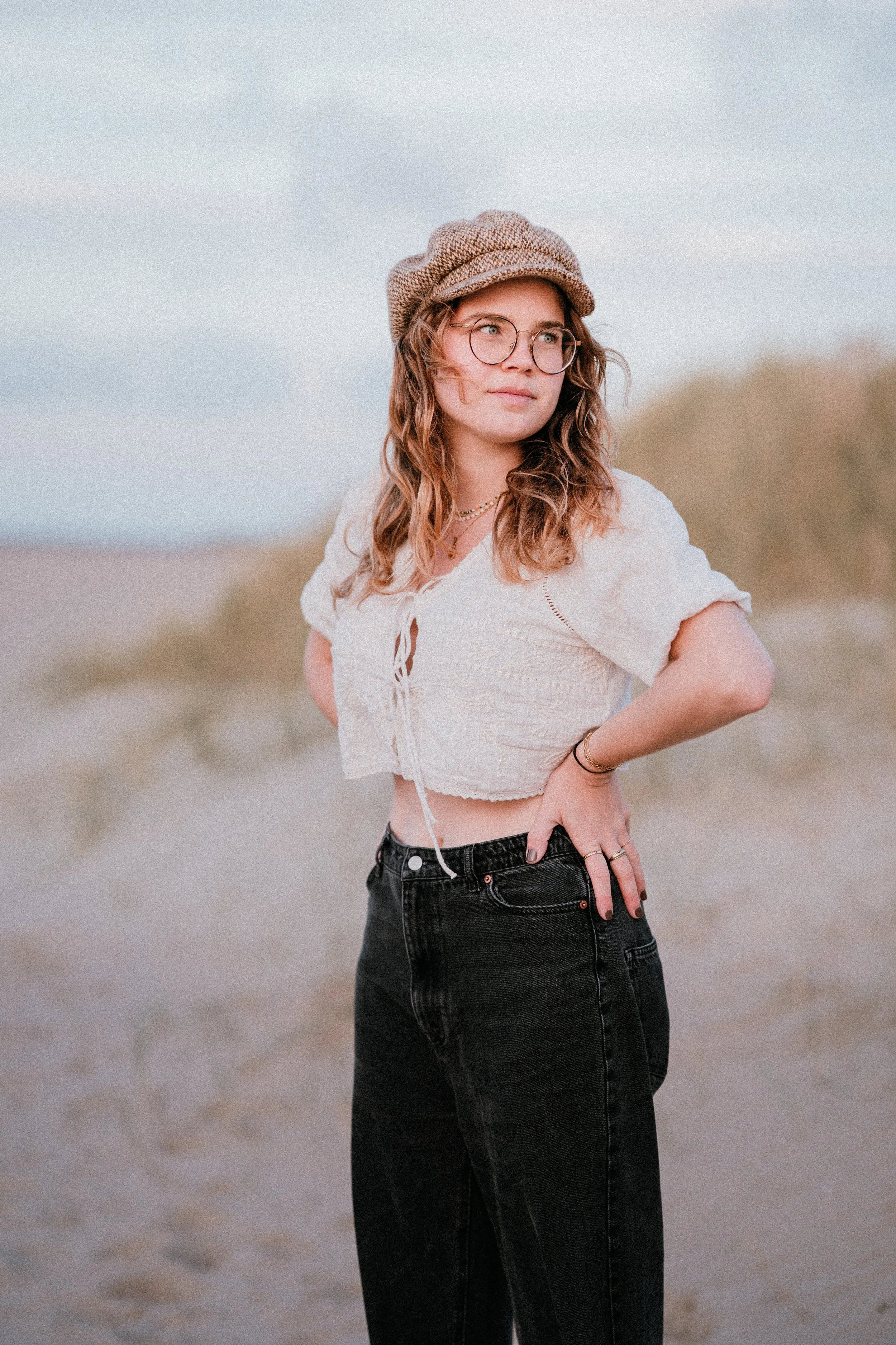 Frau mit Brille, beige Mütze, weißen Top und schwarzen Hosen steht am Strand.