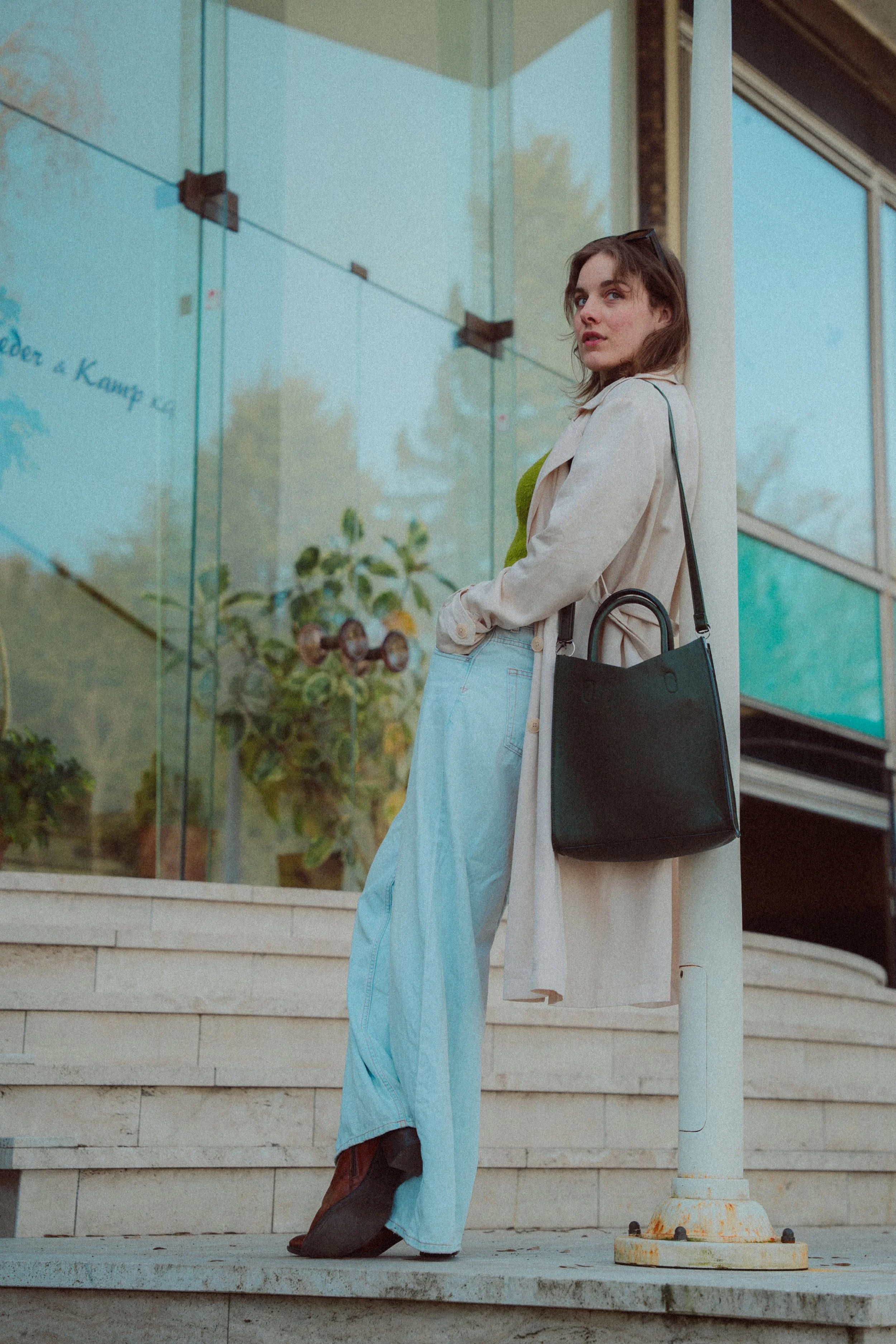 Junge Frau steht an einer transparenter Glasfensterwand, trägt beige Trenchcoat, grüne Weste, weite Jeanshose, schwarze Schuhe und hat eine schwarze Tasche. Sie schaut in die Kamera. Hinter ihr sind Pflanzen und das Gebäude ist modern gestaltet.