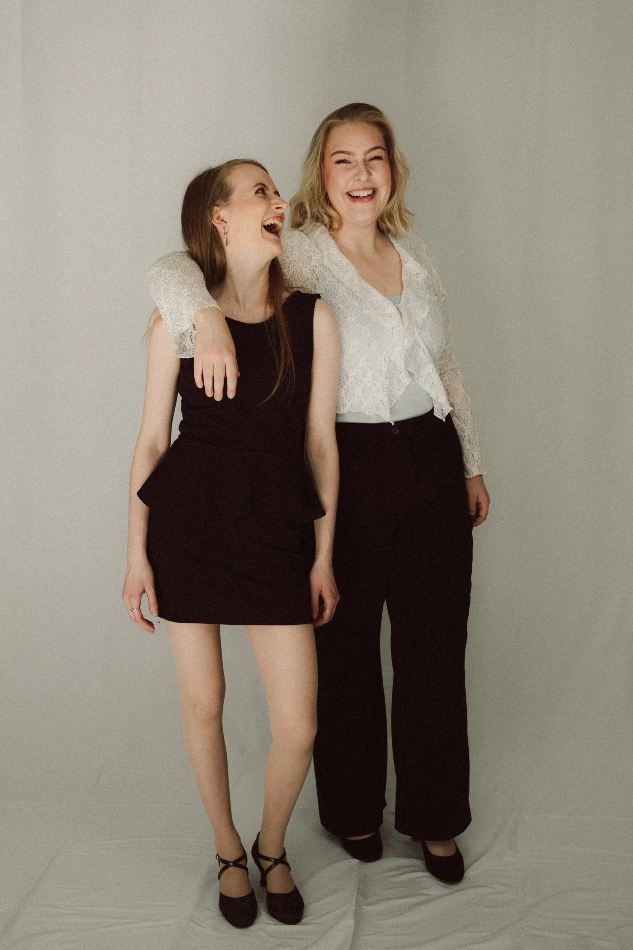Zwei Frauen, eine trägt ein schwarzes Kleid, die andere eine weiße Bluse, lachen und stehen nebeneinander vor einer neutralen Wand.