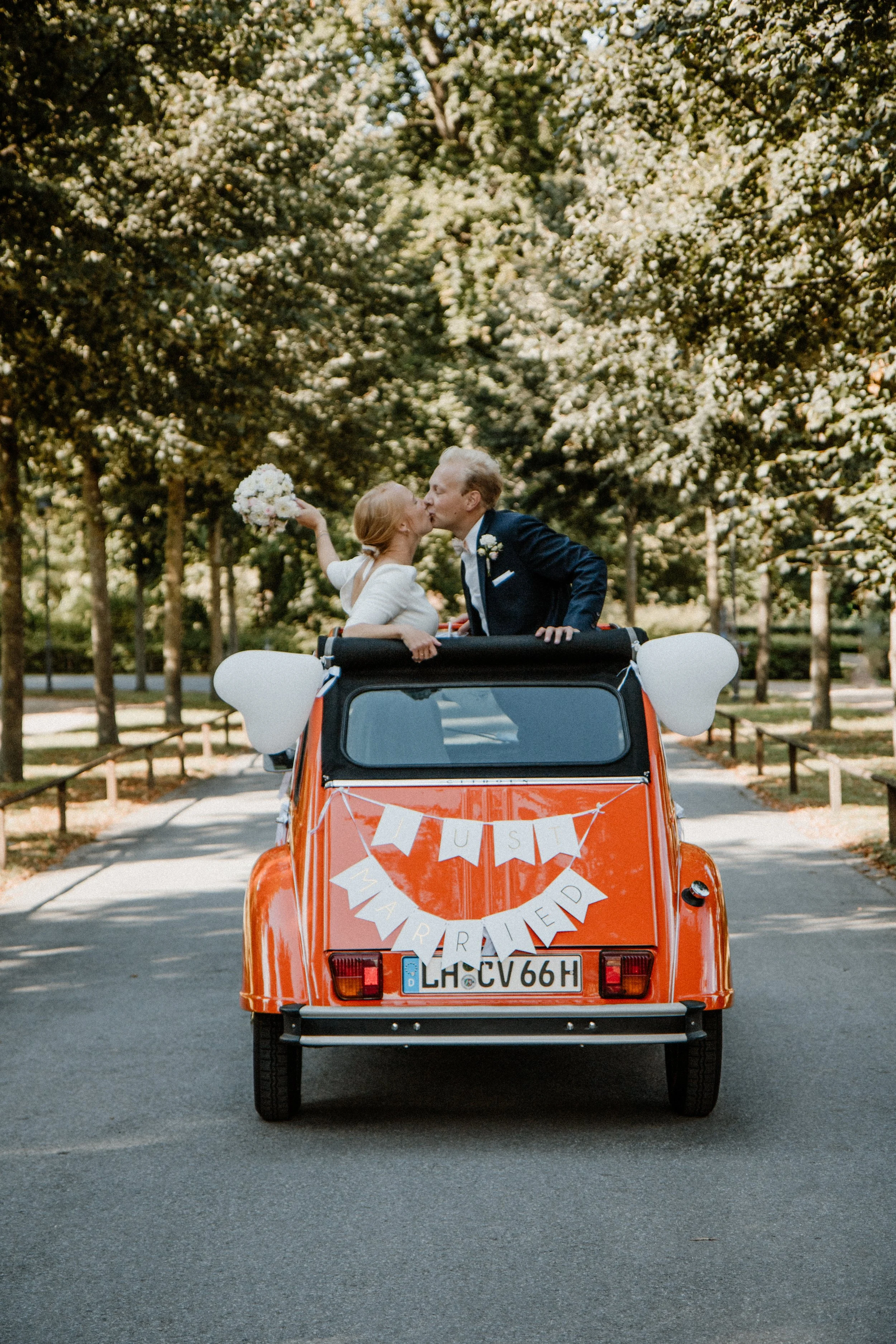 Ein rotes Auto mit weißen Herzballons an den Seiten fährt auf einer Straße. Auf der Rückseite des Autos hängt eine Fahne mit dem Schriftzug 'JUST MARRIED'. Zwei Menschen, ein Mann und eine Frau, küssen sich oben auf dem Auto. Der Hintergrund ist ein parkähnlicher Bereich mit vielen Bäumen.