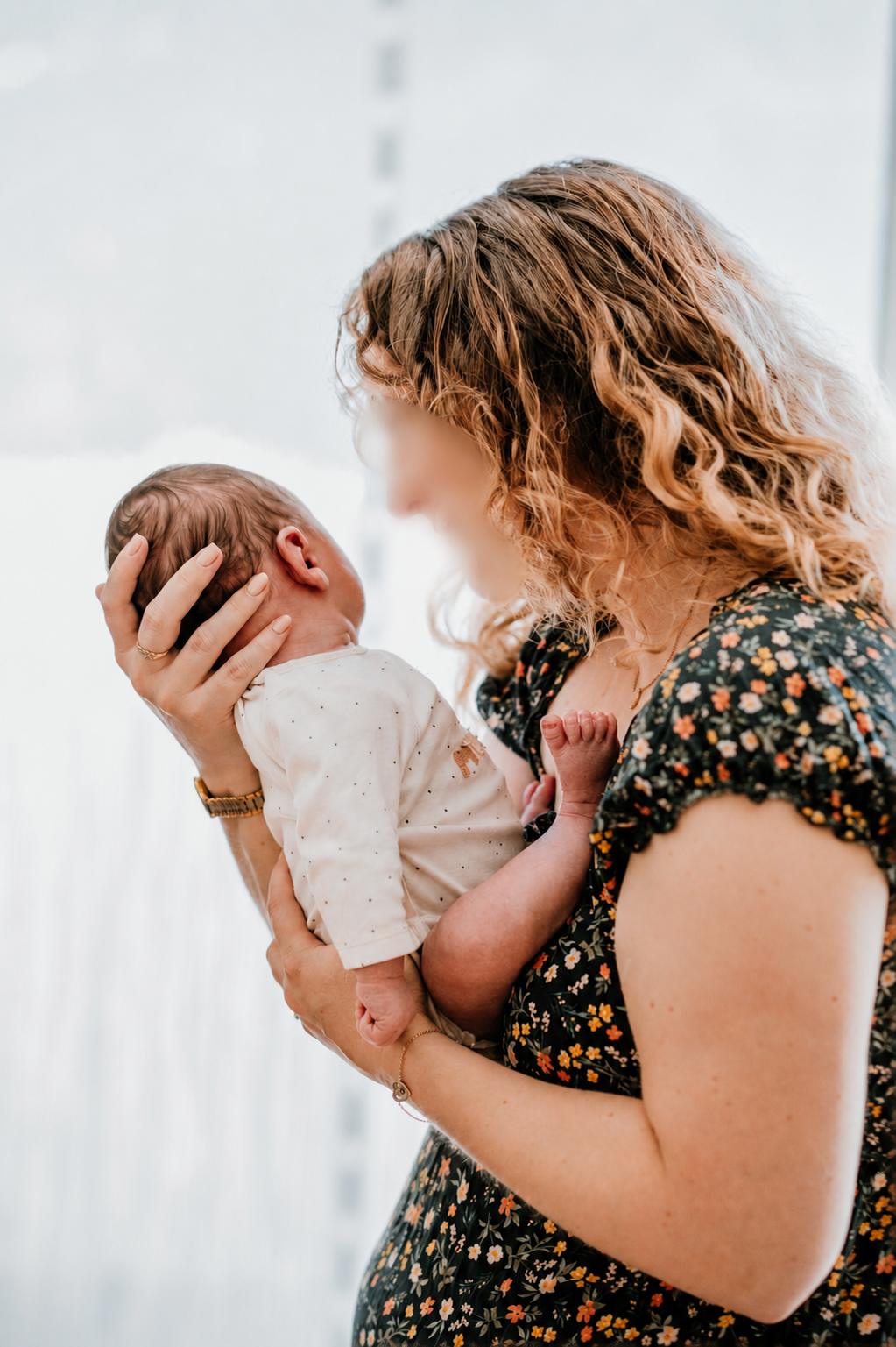 Eine junge Frau hält ein neugeborenes Baby liebevoll in den Händen, während sie es sanft an ihr Gesicht hält. Das Baby trägt ein weißes Oberteil mit kleinen Punkten.