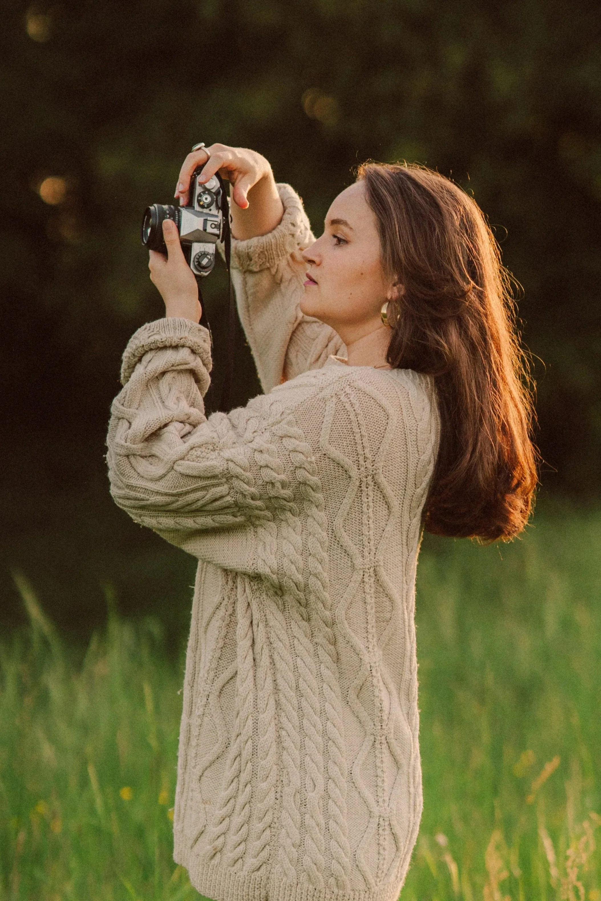 Junge Frau in einem cremefarbenen, strickgeknüpften Kleid steht in einem grünen Feld bei Sonnenuntergang und fotografiert mit einer Kamera.
