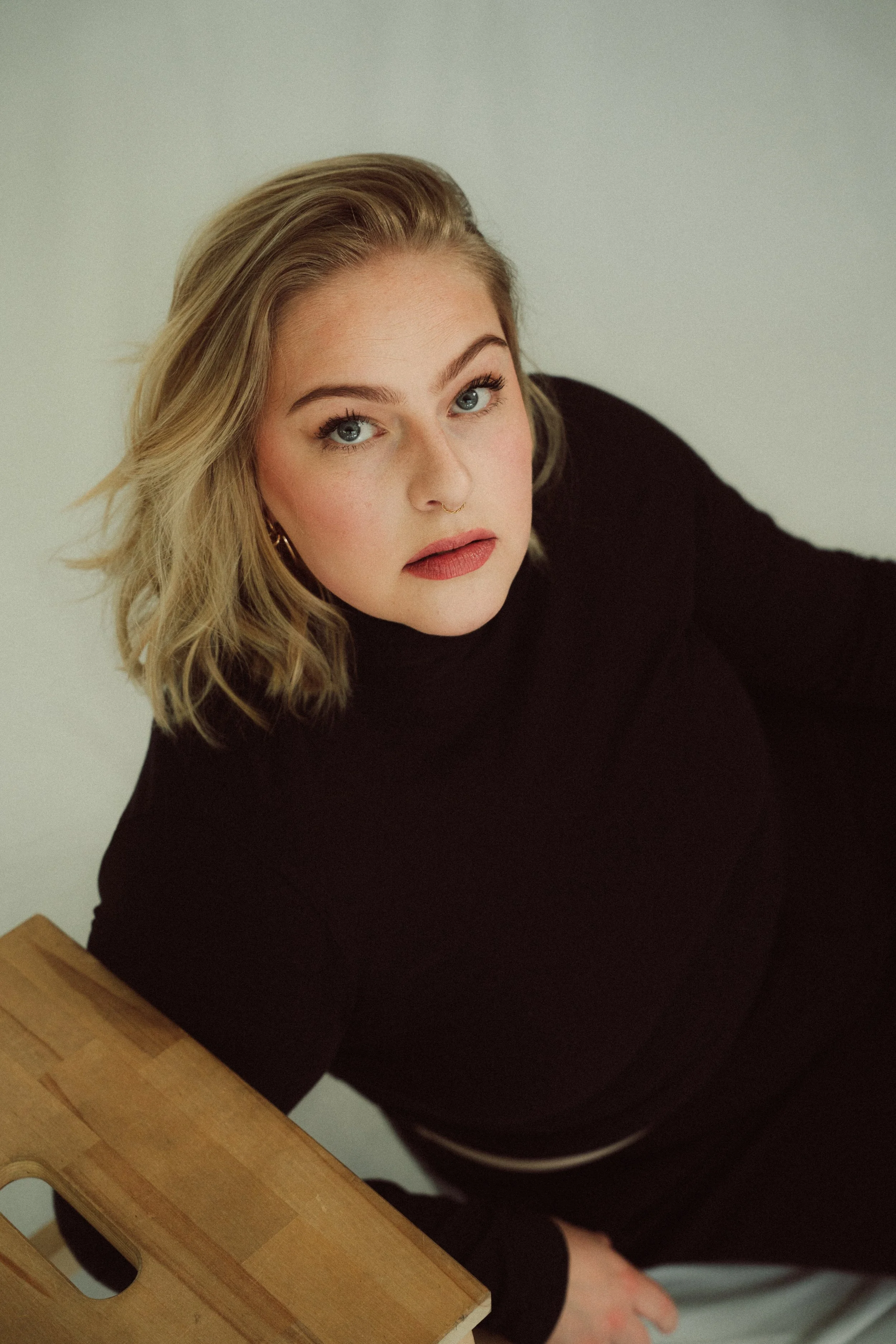 Nahaufnahme eines jungen Mädchens mit blonden, lockigen Haaren, blauen Augen und dezentem Make-up, trägt ein schwarzes Oberteil und sitzt an einem Holztisch.