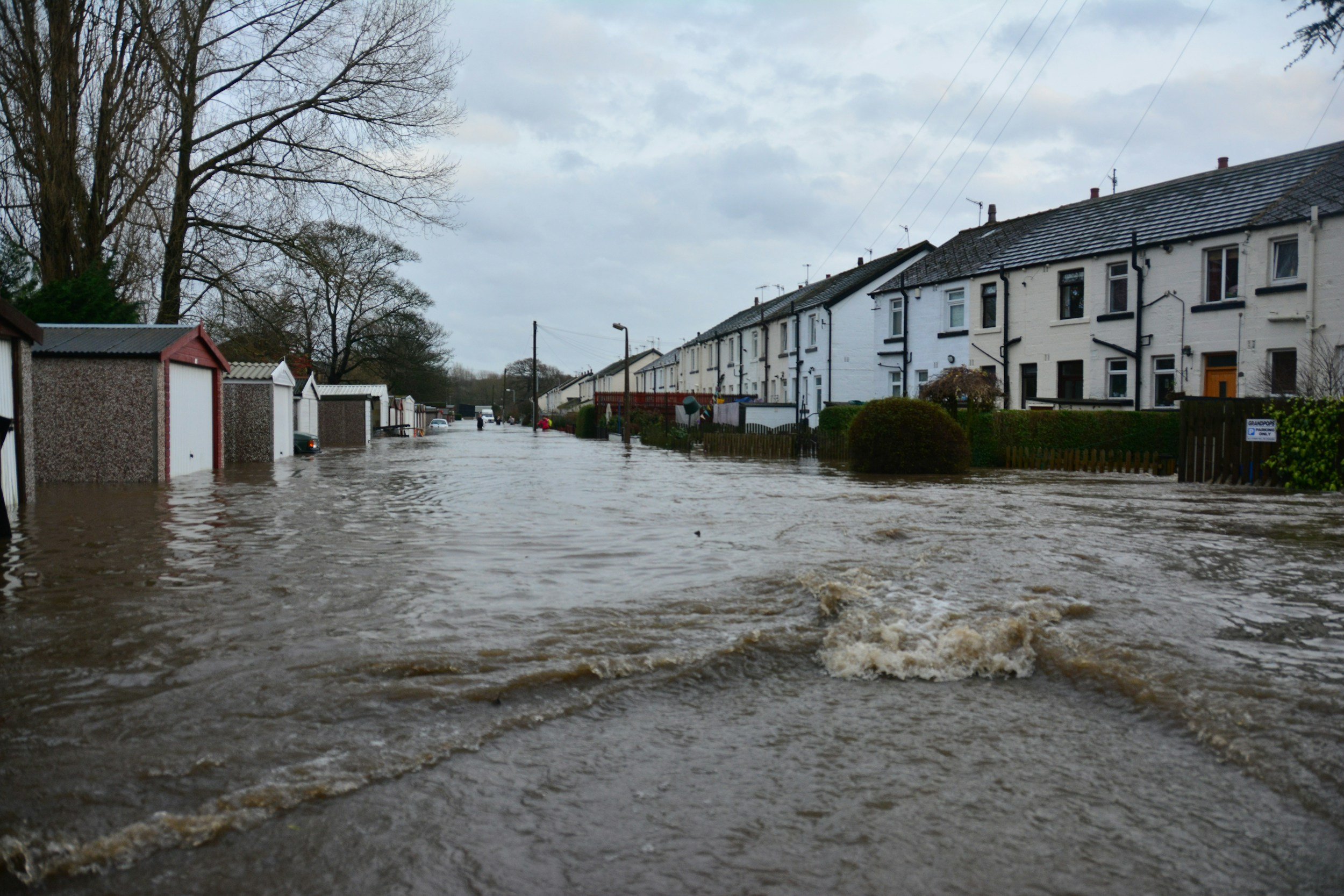 Flooding on a residential street with water covering the road and partially submerging parked cars. Row of white houses with rooftops and trees in the background under a cloudy sky.