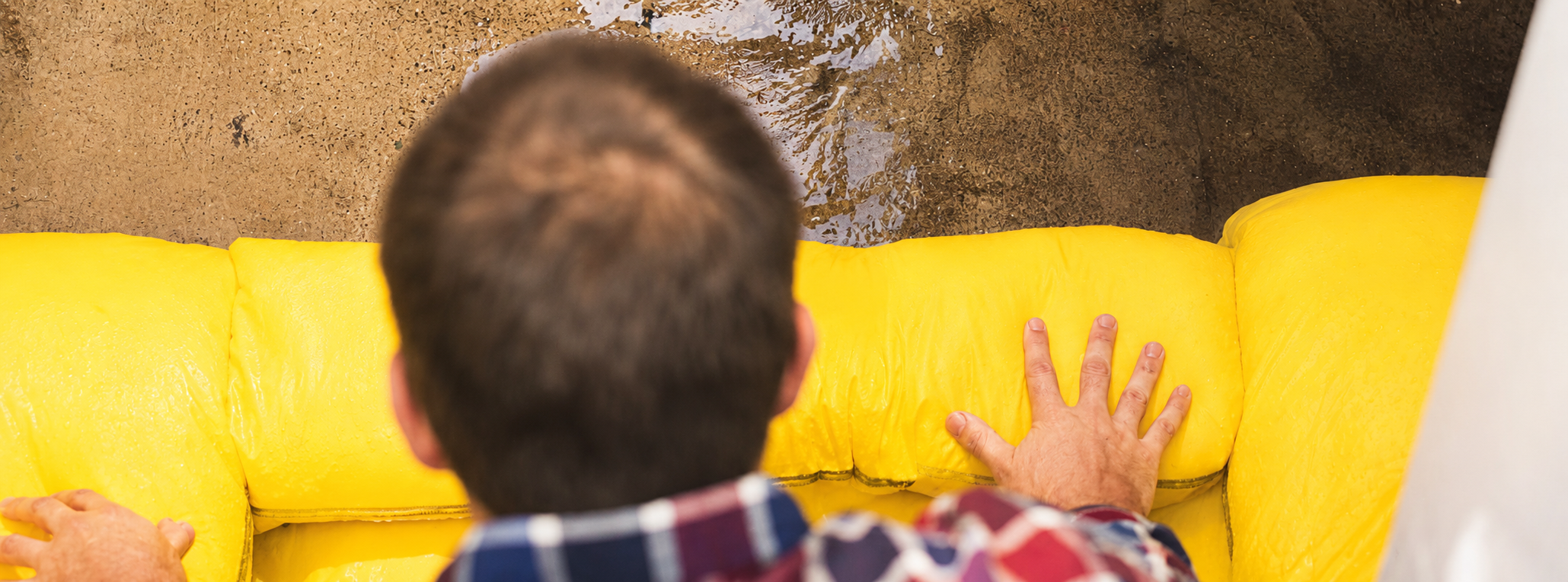 A person with short brown hair and wearing a plaid shirt touches a yellow couch with their left hand, seen from behind, with a wet concrete floor above.