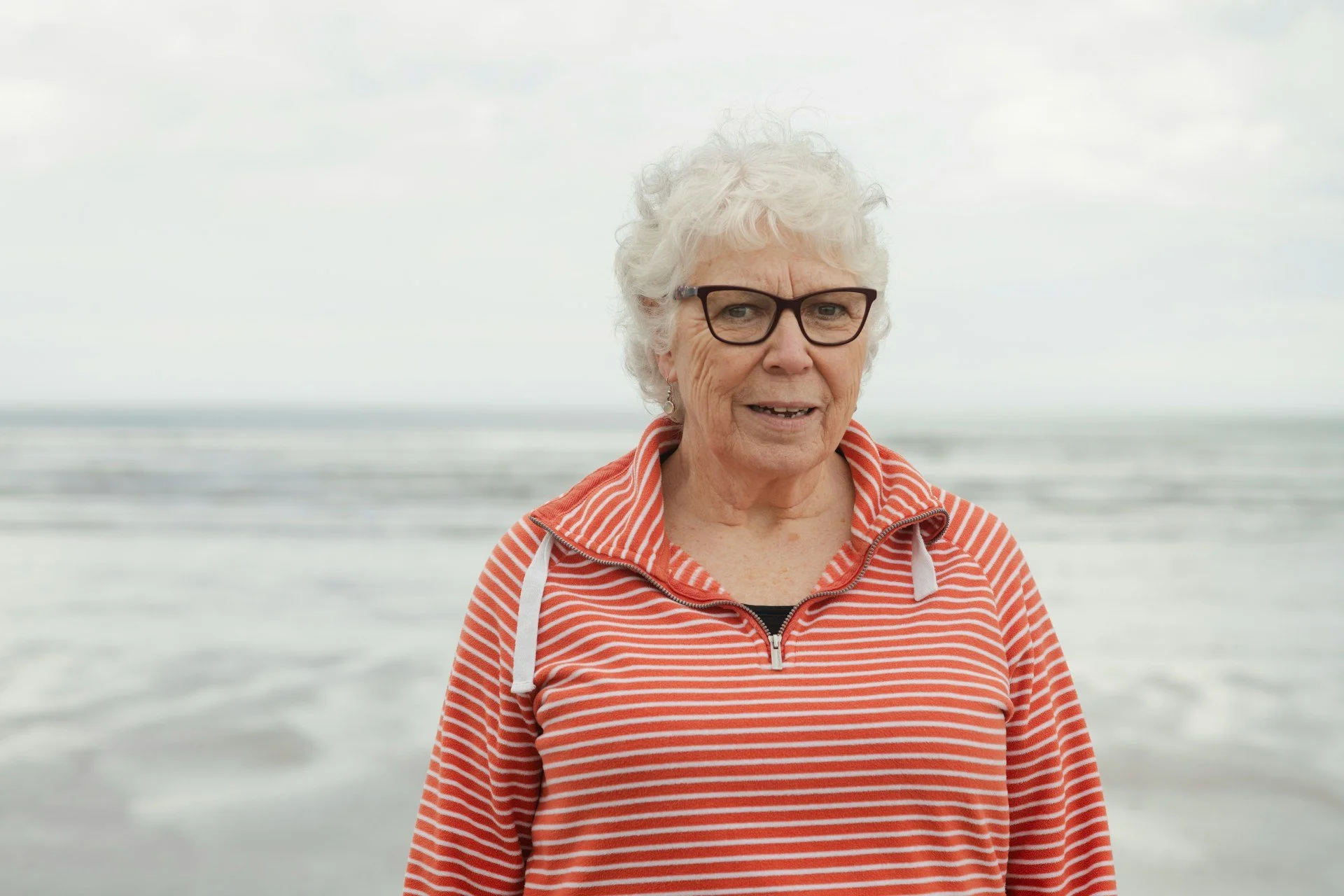 An elderly woman with curly gray hair, wearing glasses and a red and white striped hoodie, standing on a beach with waves in the background.