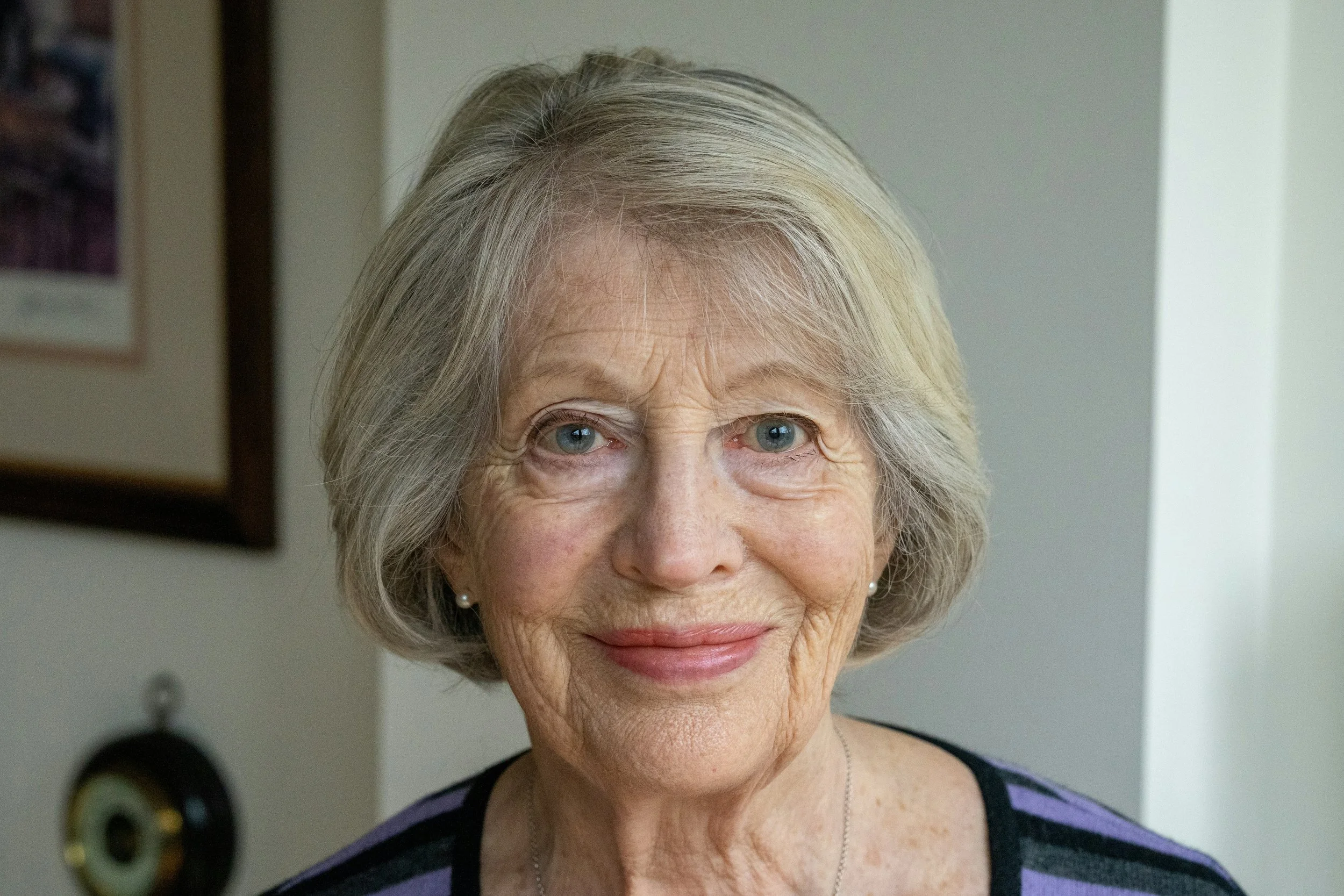 Close-up of an elderly woman with short gray hair, blue eyes, and a gentle smile, wearing pearl earrings and a black and purple striped top, in a room with framed pictures in the background.