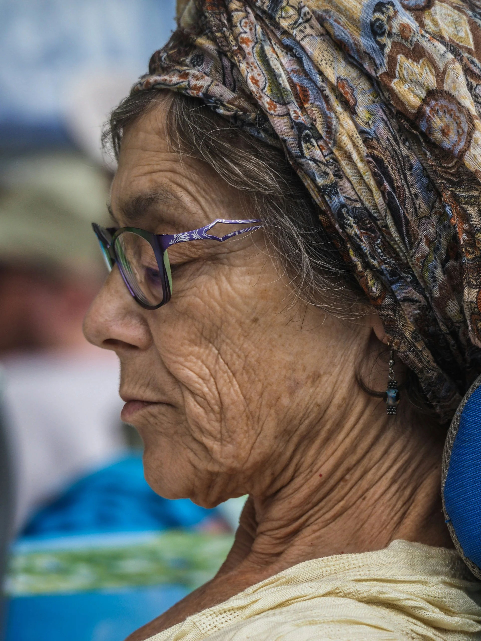 A close-up side profile of an elderly woman with glasses and a patterned headscarf, showing facial wrinkles and ear jewelry.