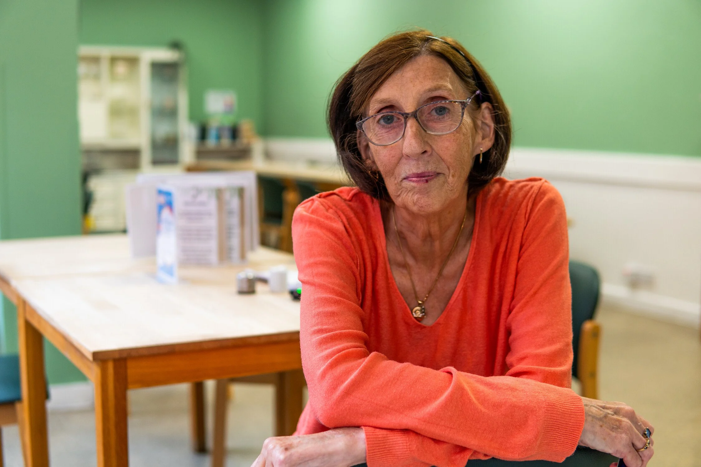 Older woman with brown hair, glasses, and earrings sitting at a table in a green-painted room, wearing an orange sweater.