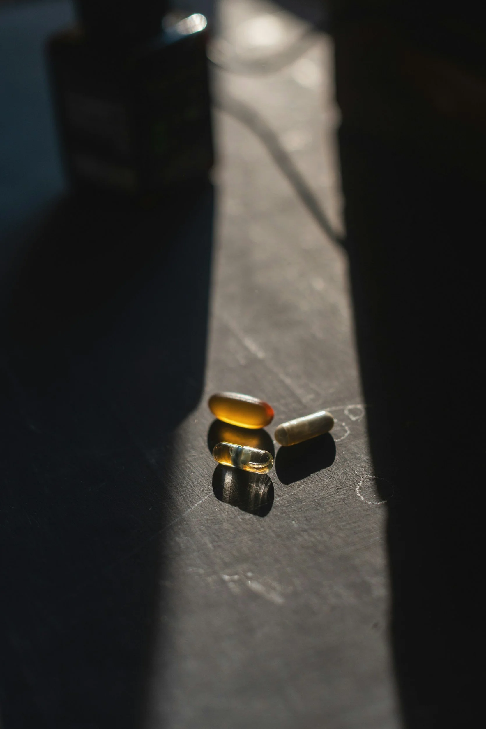 Close-up of a few scattered capsules and pills on a dark surface with sunlight casting shadows.