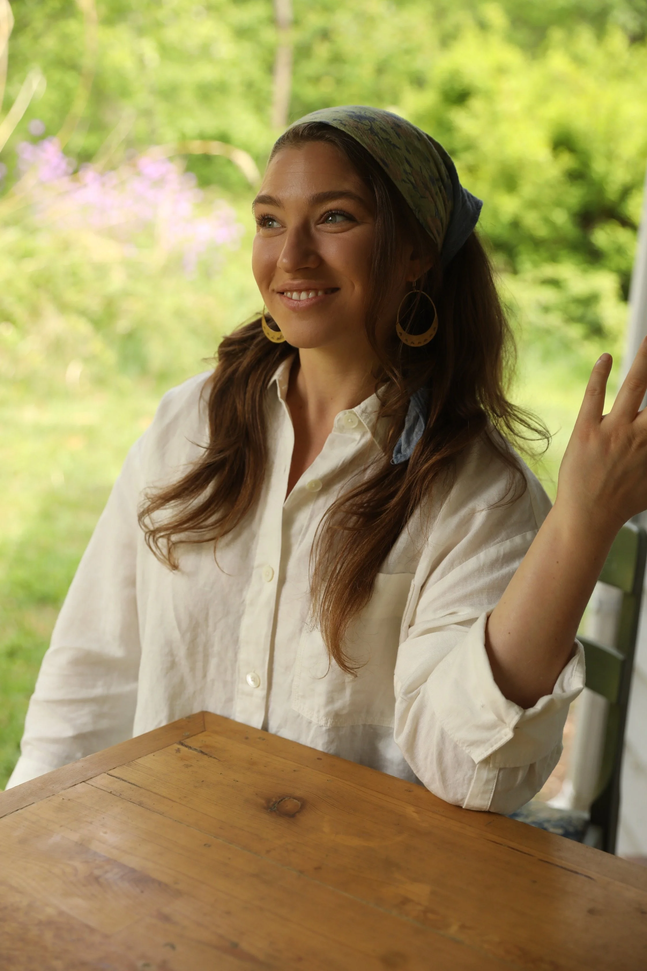 A young woman sitting at a wooden table, smiling, with a green leafy background, wearing a white shirt, large hoop earrings, and a headscarf.