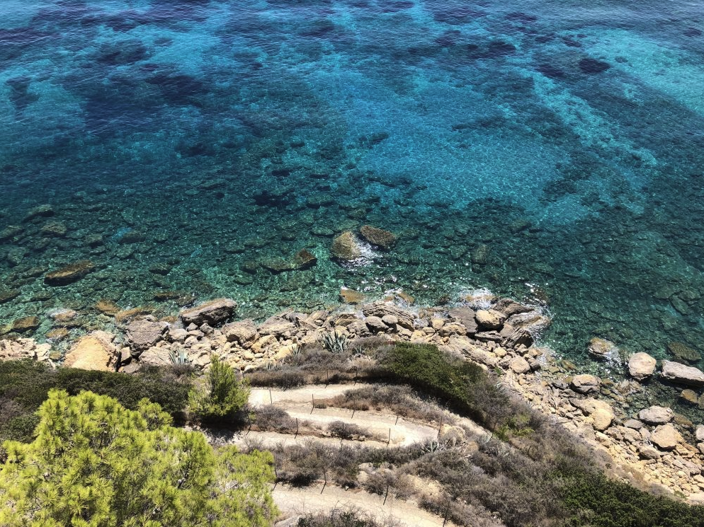 Aerial view of a rocky shoreline along a clear blue body of water, with sparse coastal vegetation and a dirt path.