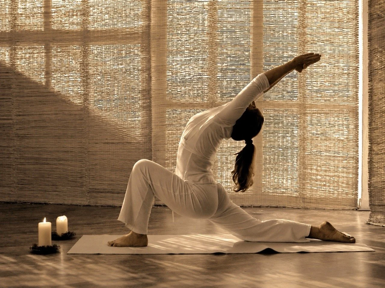 A woman practicing yoga indoors on a mat, performing a side stretch pose with candles nearby, in front of woven blinds that let sunlight through.