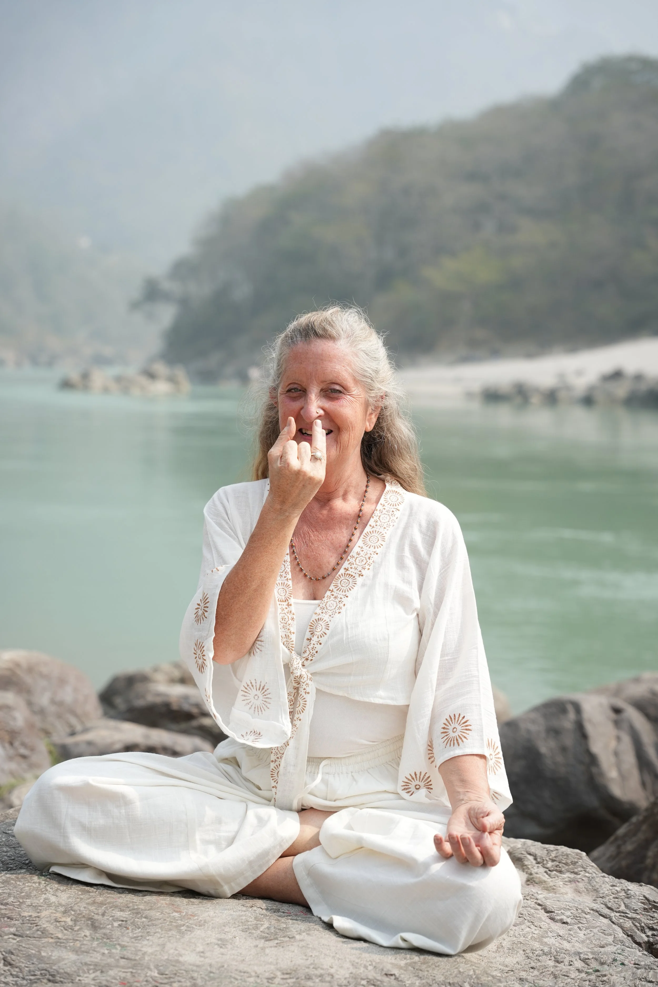 An elderly woman with gray hair tied in a bun, sitting cross-legged on some rocks by the water, practicing yoga or meditation. She is smiling and plays with her nose with her index finger. She is dressed in a white, embroidered, flowing outfit. The background features a body of water and hills covered with trees.