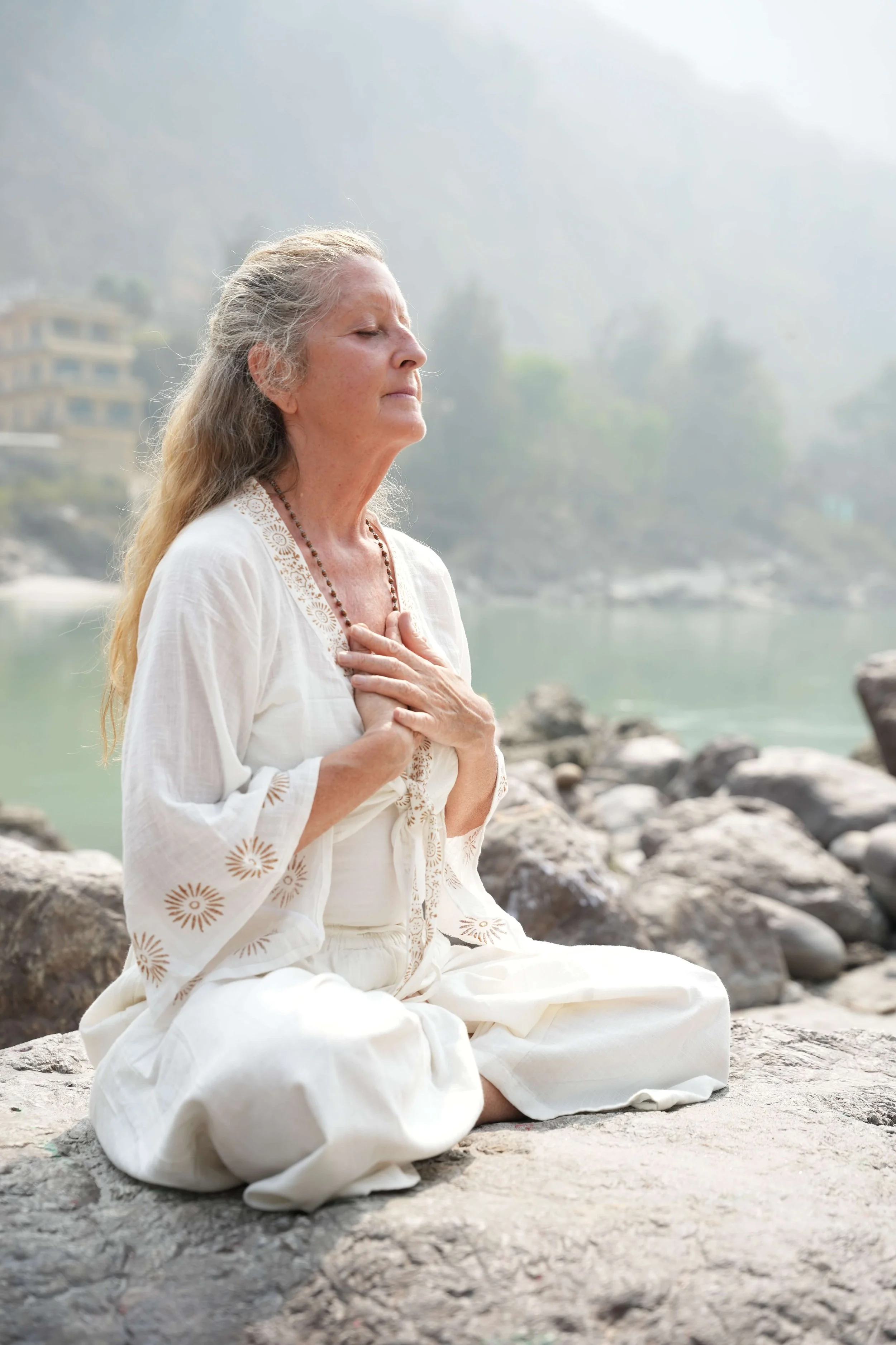 An elderly woman with gray hair, dressed in white, is sitting on rocky ground near a body of water with rocks, her eyes closed, hands over her chest, appearing to meditate or pray in a serene outdoor setting with misty mountains and trees in the background.
