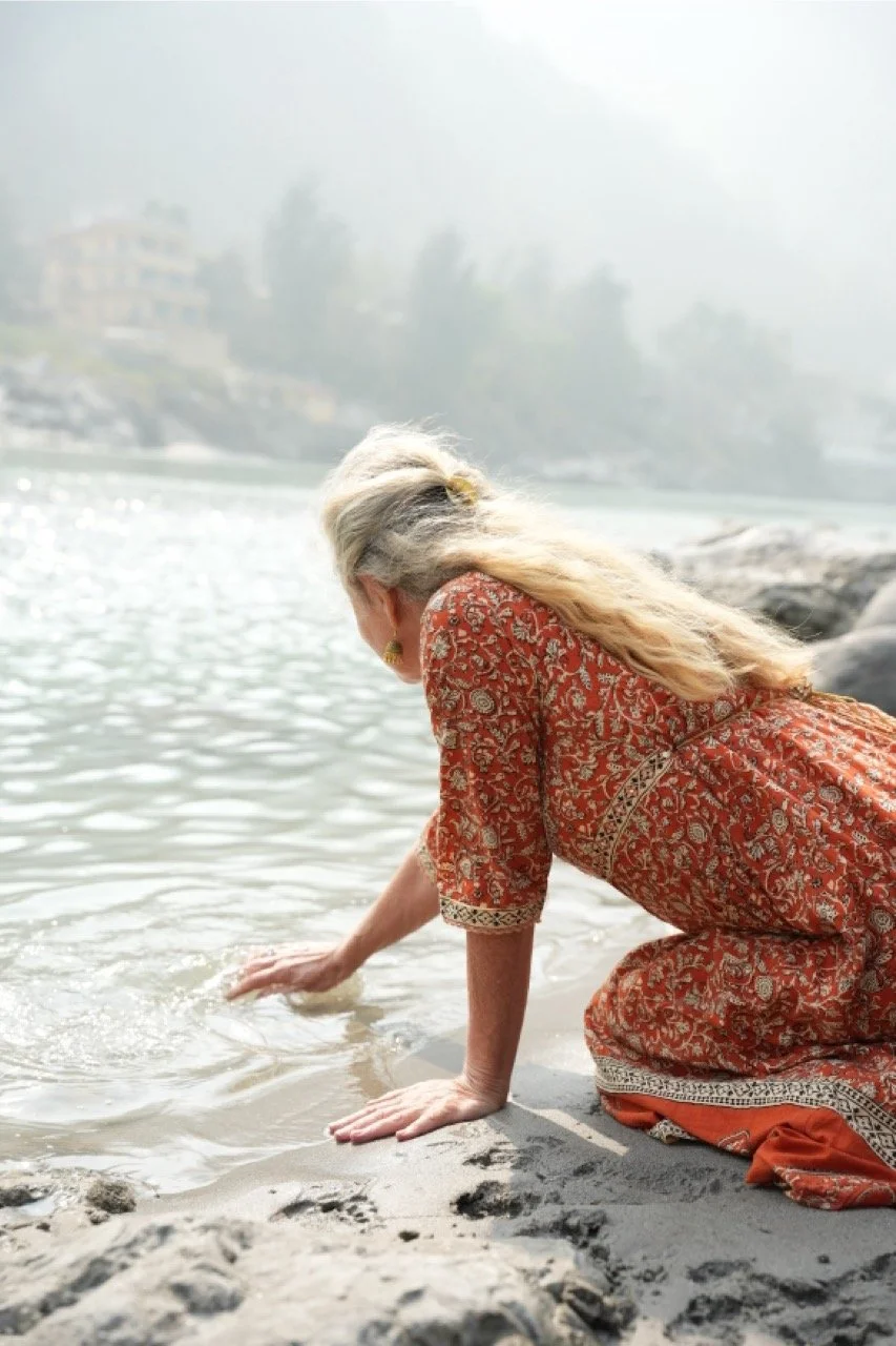 An older woman in a red patterned dress with gold accents kneeling on sandy beach, reaching into the water with one hand.
