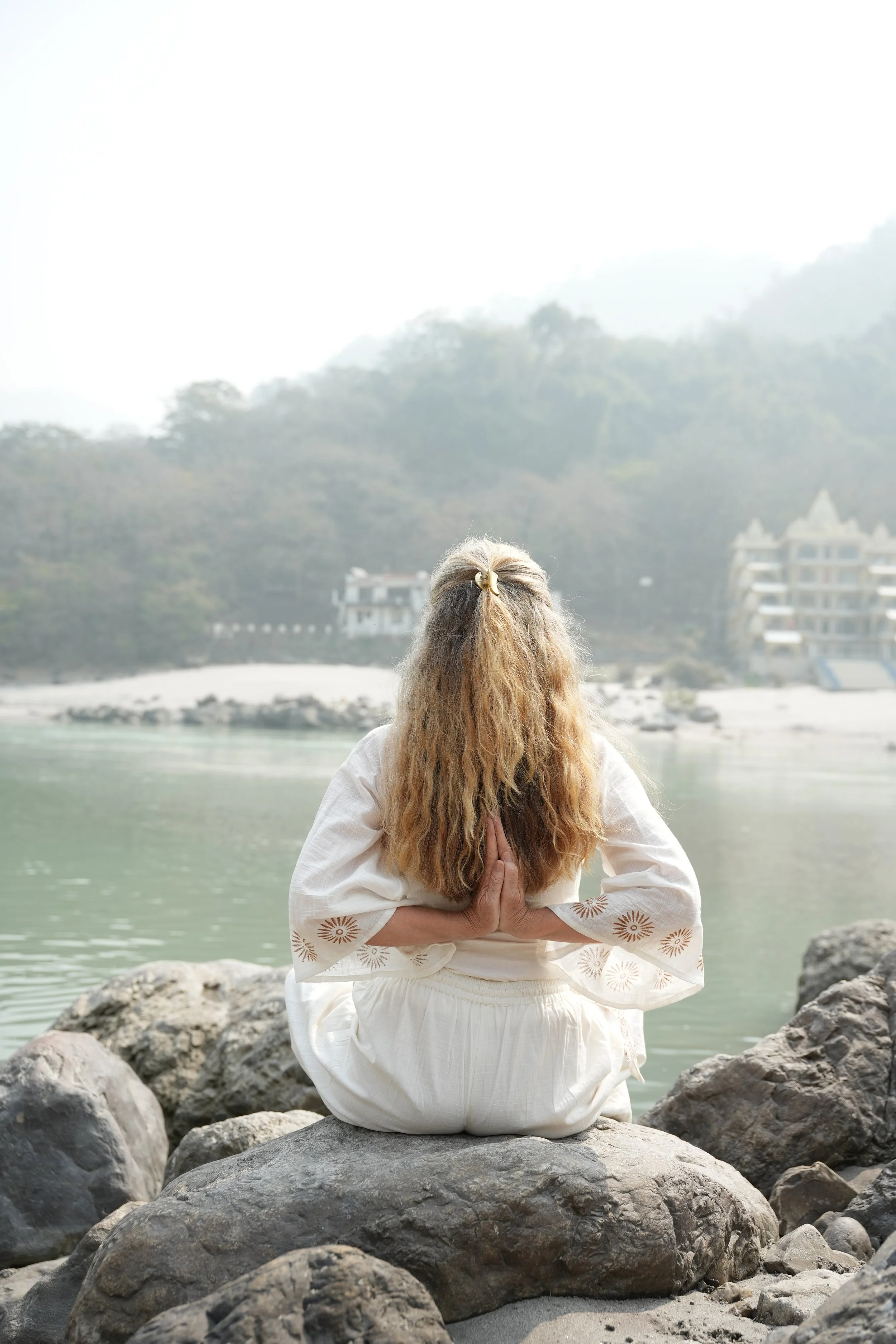 A woman with long curly hair in a white dress practicing yoga on a rocky beach with a body of water and trees in the background.