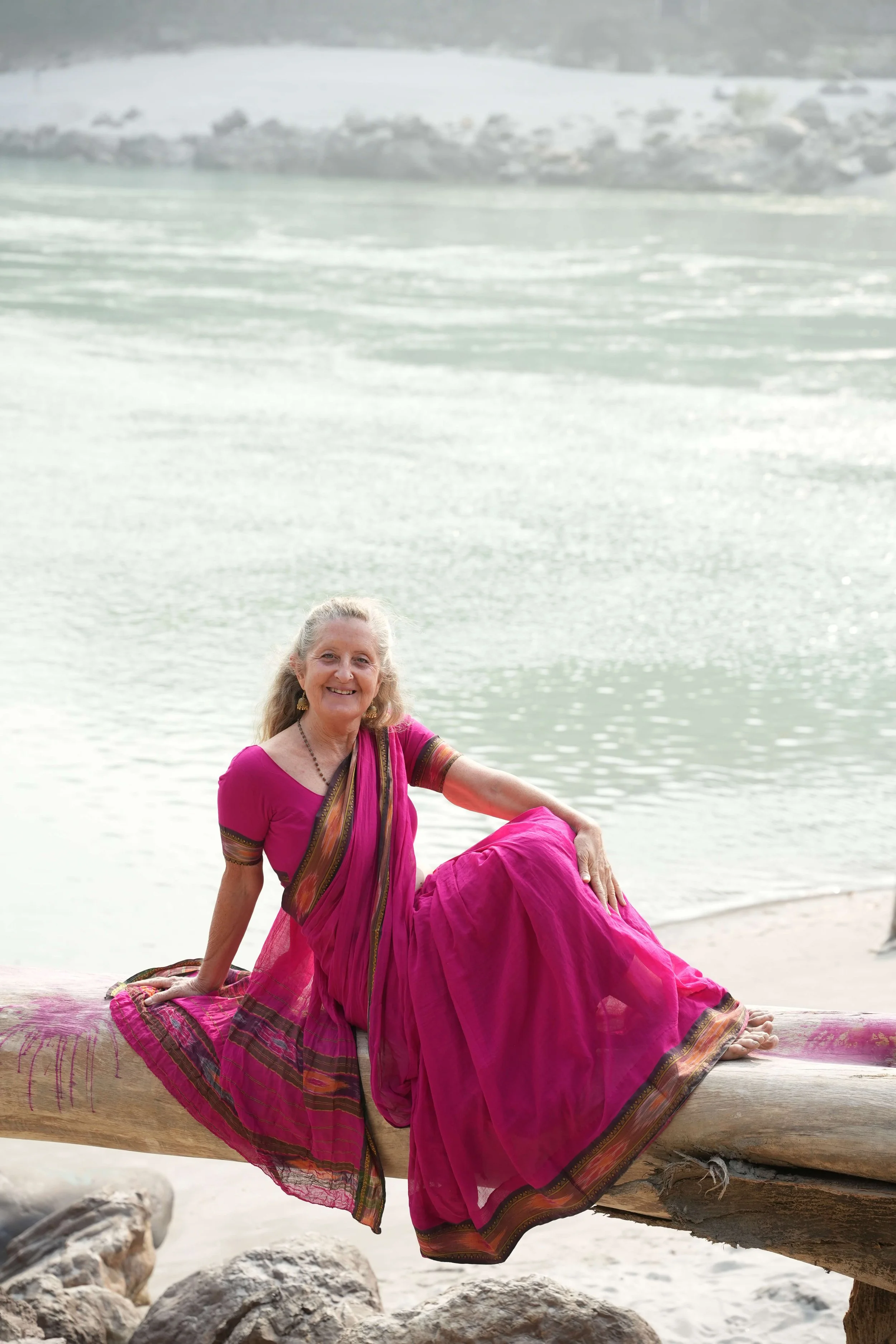 A woman with gray hair wearing a pink traditional Indian sari, sitting on a large wooden log near a body of water with rocks around, smiling at the camera.