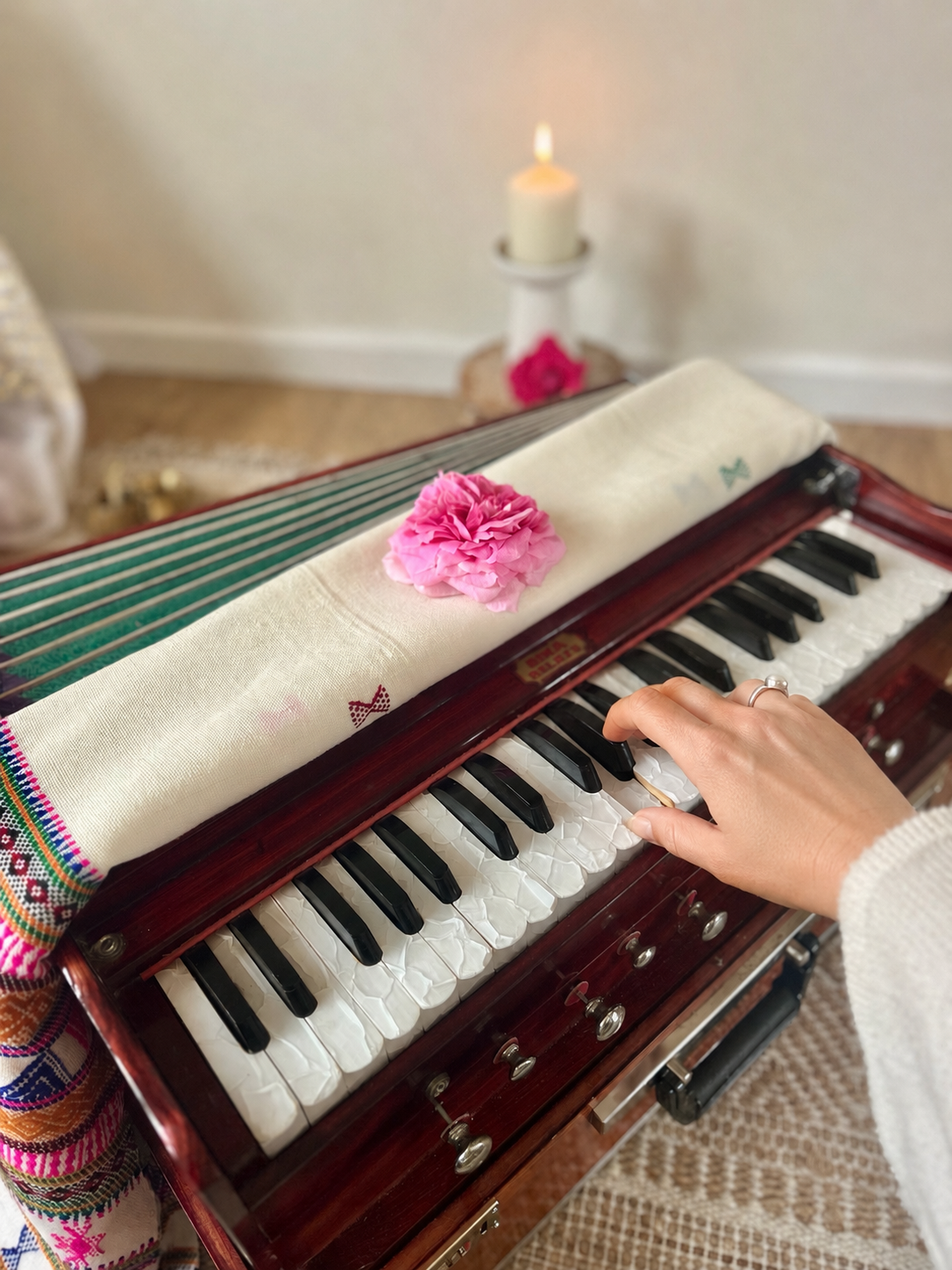 A person playing a small vintage wooden piano with a pink flower on the cover, a white cloth draped over it, and a pink flower in the background on a candle holder.