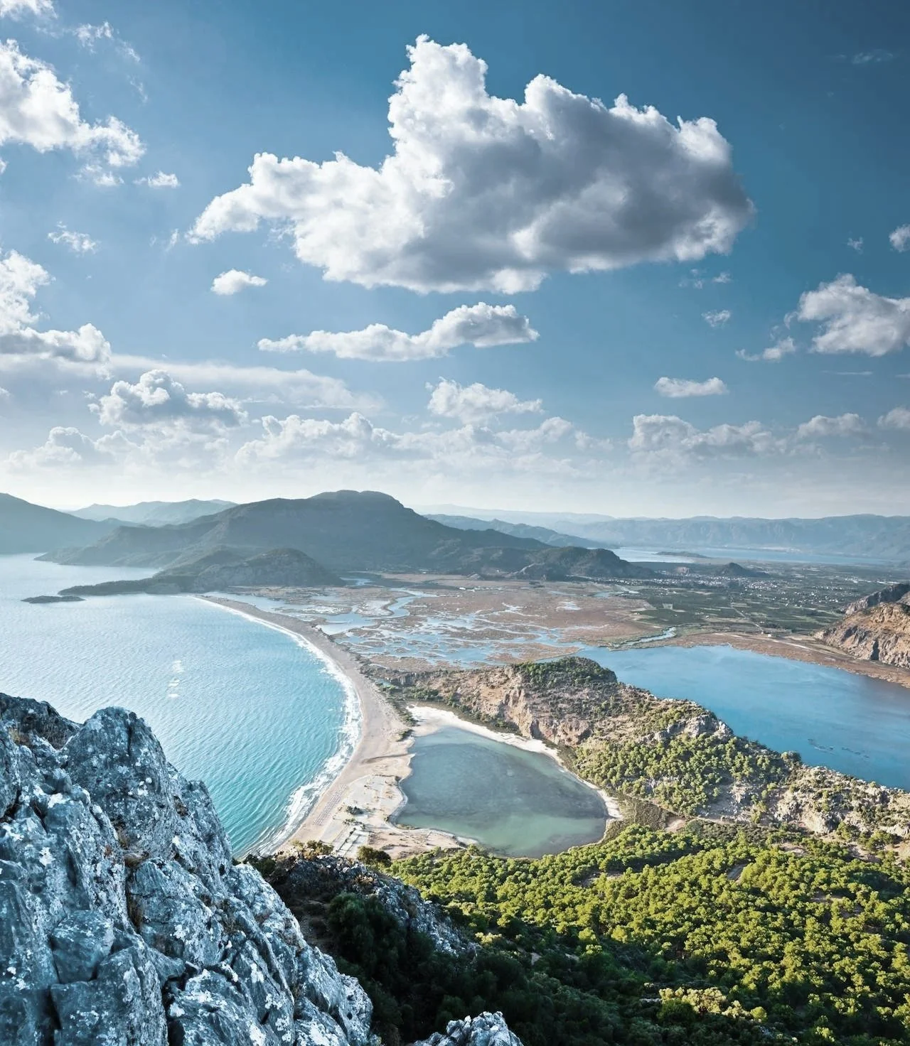 A panoramic view of a river delta with a sandy shoreline, surrounded by mountains and lush green forests under a partly cloudy sky.
