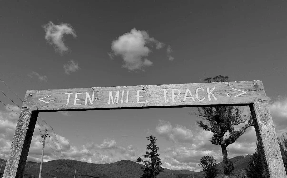 Wooden sign indicating the 'Ten Mile Track' with arrows pointing left and right, against a background of mountains, trees, and a partly cloudy sky.