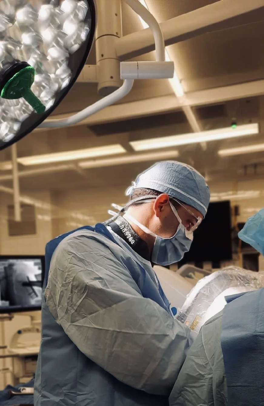 A surgeon dressed in scrubs, mask, and protective glasses performing surgery in an operating room with medical equipment and bright surgical lights above.