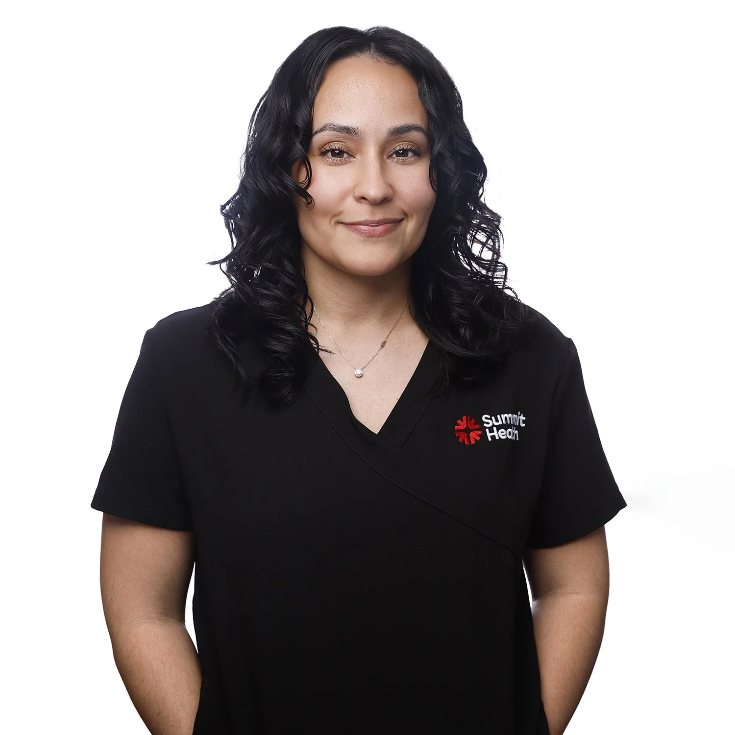 A woman with long black curly hair wearing a black medical scrub top with a Summit Health logo, smiling with her right hand in her pocket, against a white background.