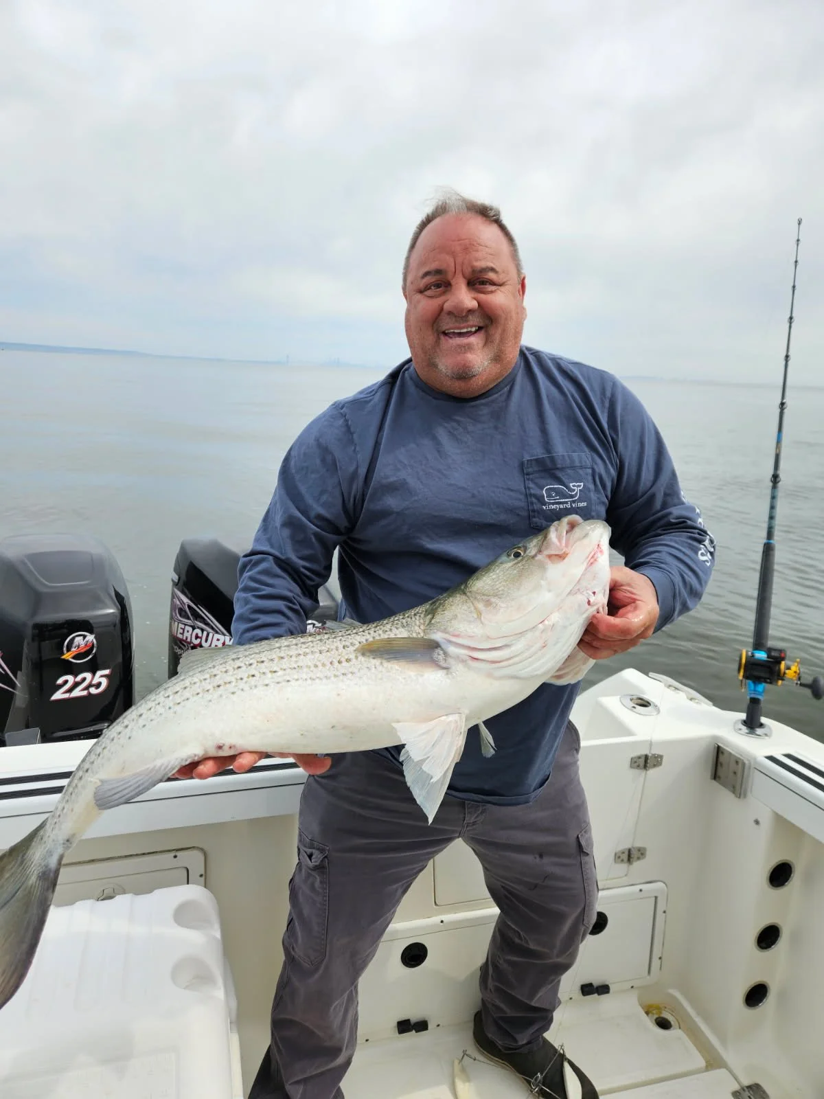 Man on boat holding a large striped bass fish with a cloudy sky and water in the background.