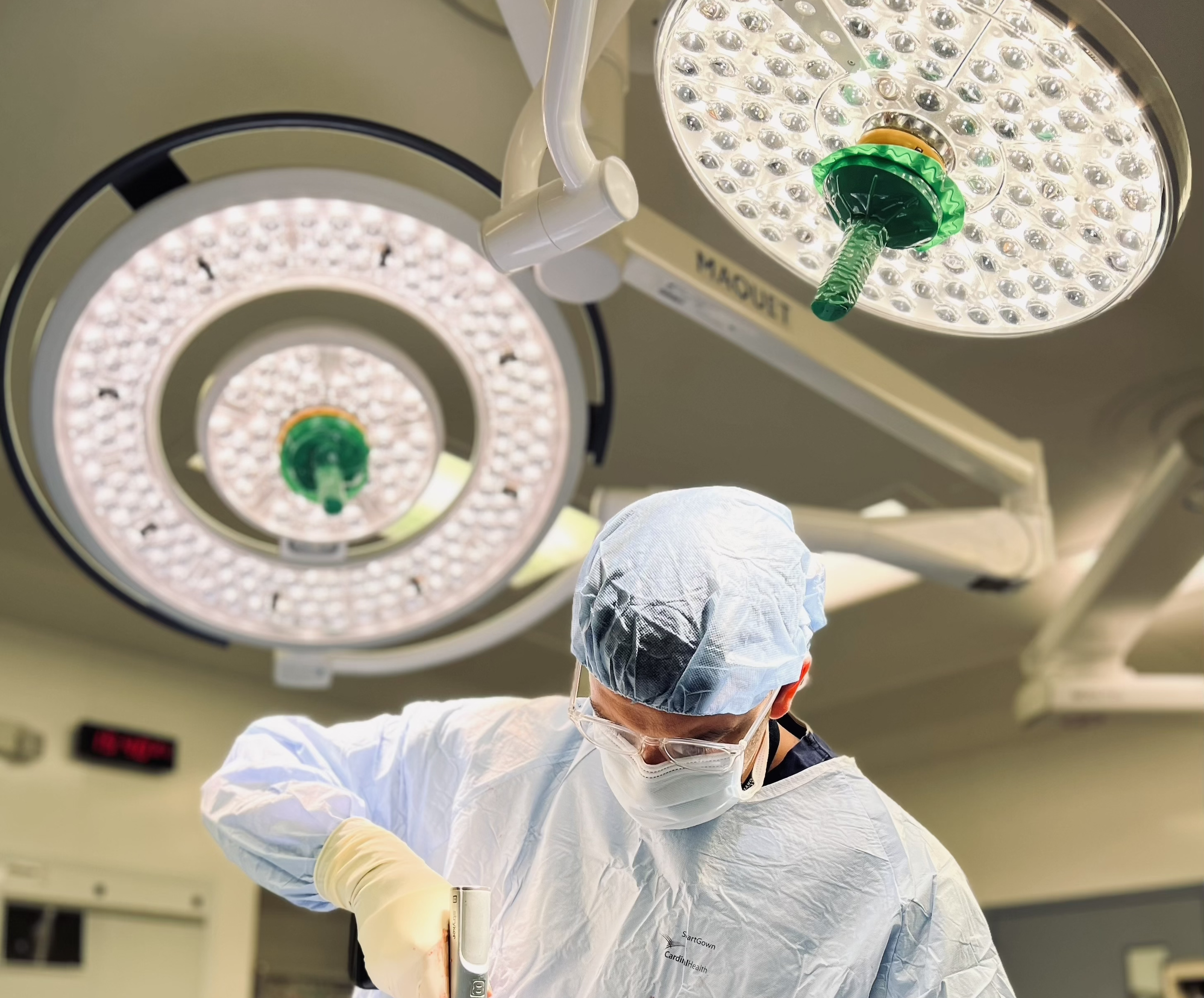 A healthcare professional wearing scrubs, a surgical cap, mask, and gloves is preparing to perform a procedure in an operating room. Overhead surgical lights are visible above.