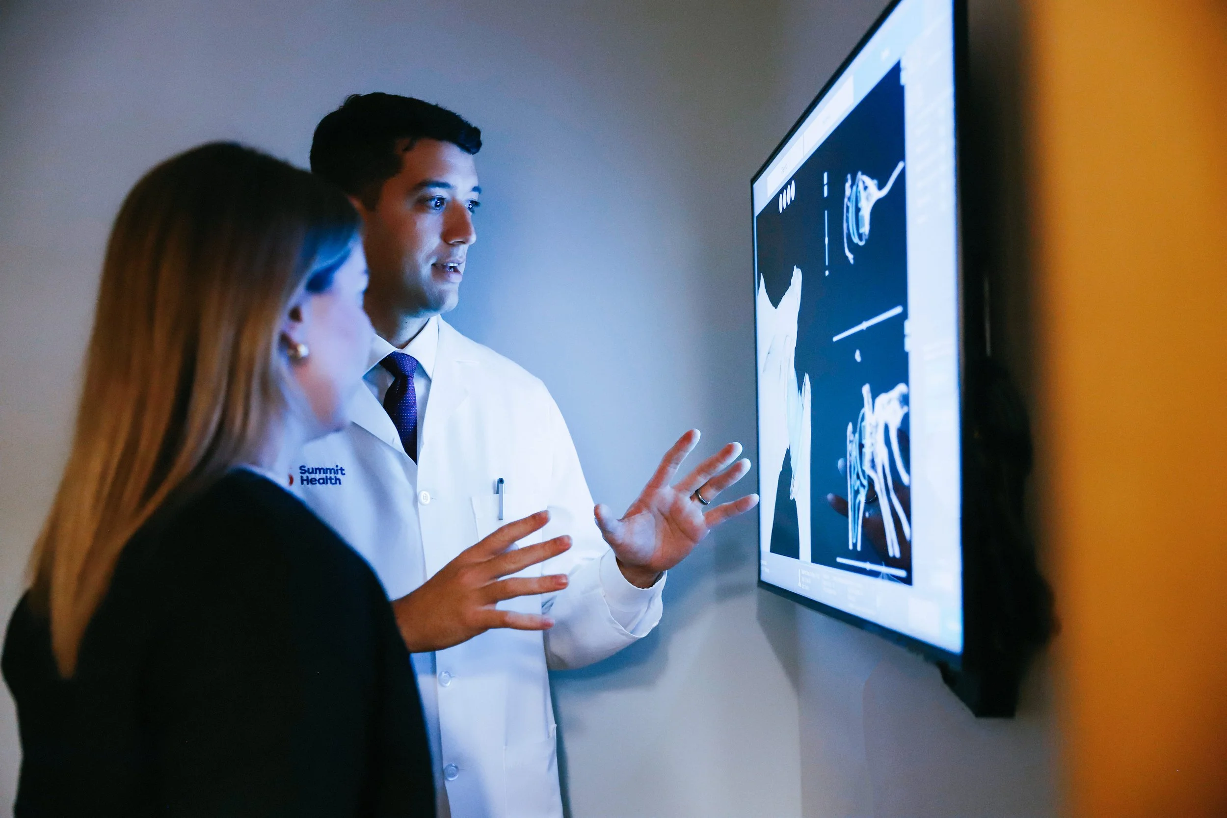 A male doctor and a female patient looking at a large monitor displaying medical images, likely MRI or CT scans, in a clinical setting.