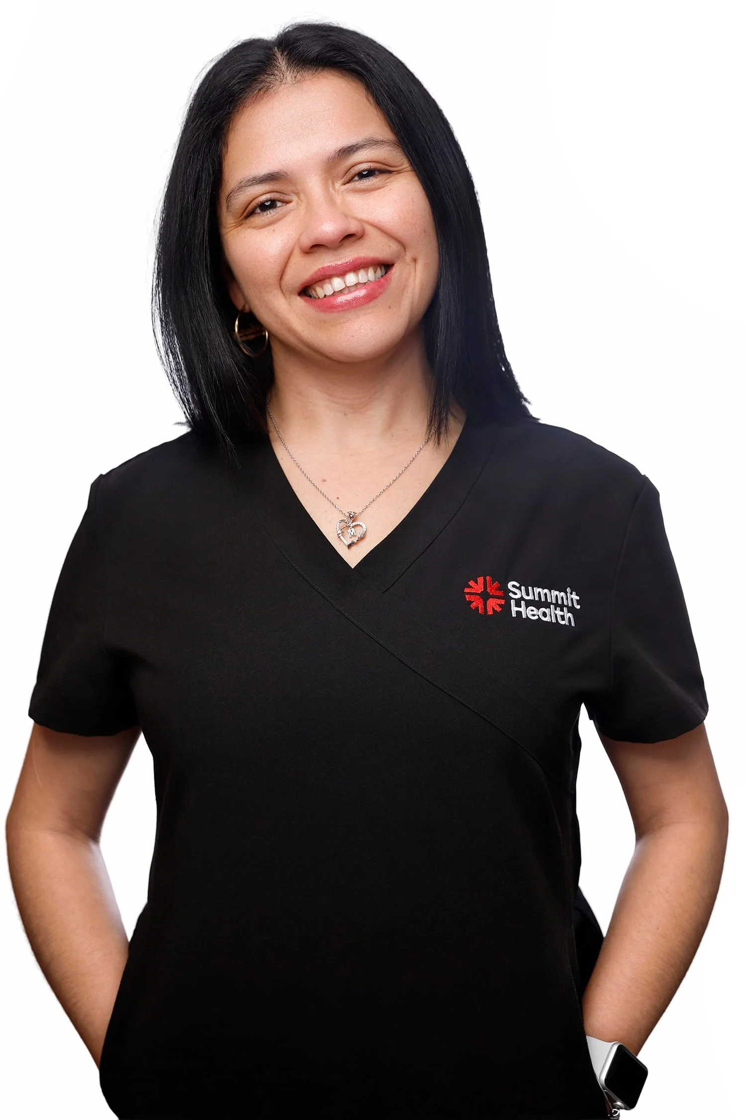 A woman with black hair smiling, wearing a black Summit Health medical uniform, a heart-shaped necklace, and a smartwatch on her left wrist, standing against a white background.