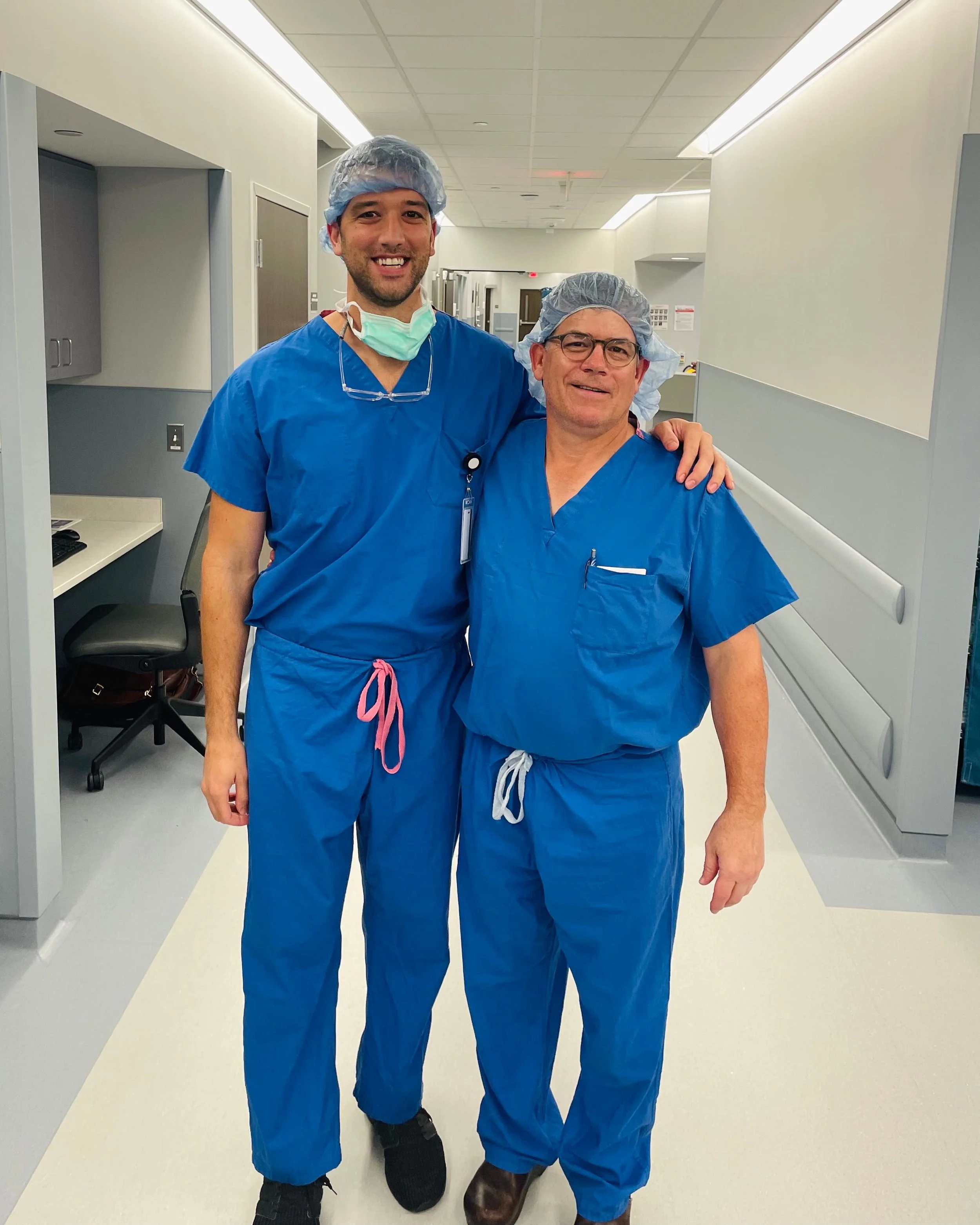 Two male healthcare professionals wearing blue scrubs, hair coverings, and face masks, standing in a hospital corridor and smiling at the camera.