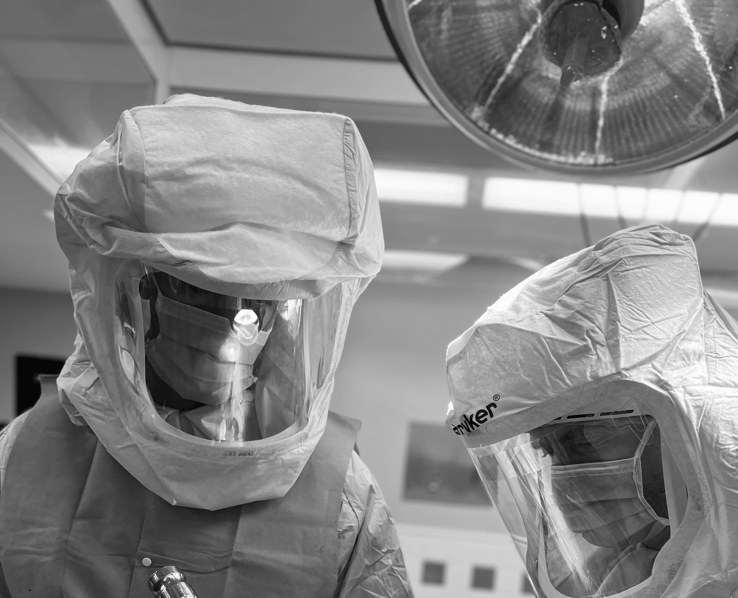 Two individuals wearing protective gear, including gloves, surgical masks, and gowns, working in a clinical or laboratory setting. Overhead surgical or examination light is visible.