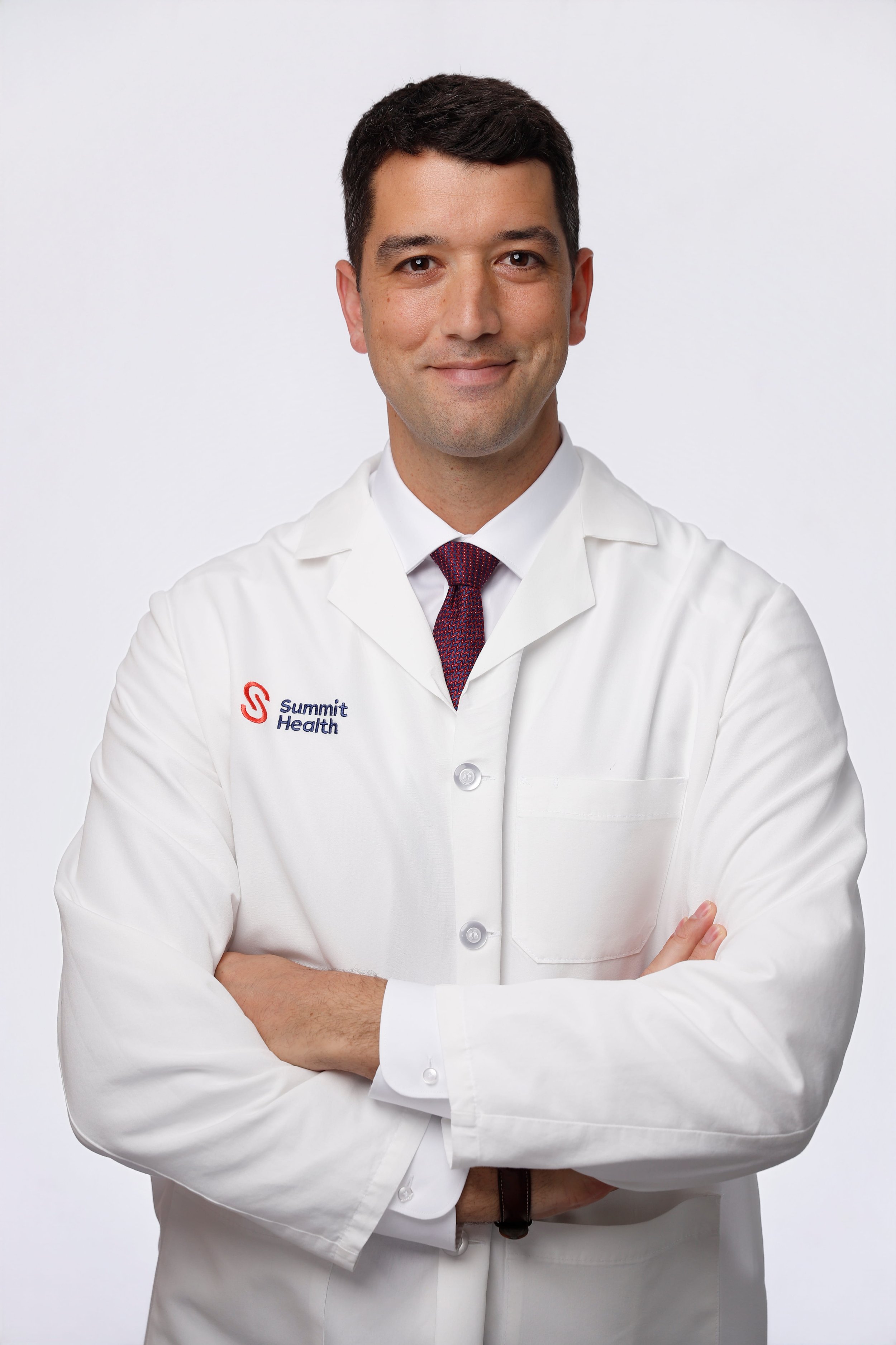 A man dressed in a white lab coat with a pink and blue tie, standing with his arms crossed, smiling at the camera. The lab coat has the logo and text "Summit Health" on it.