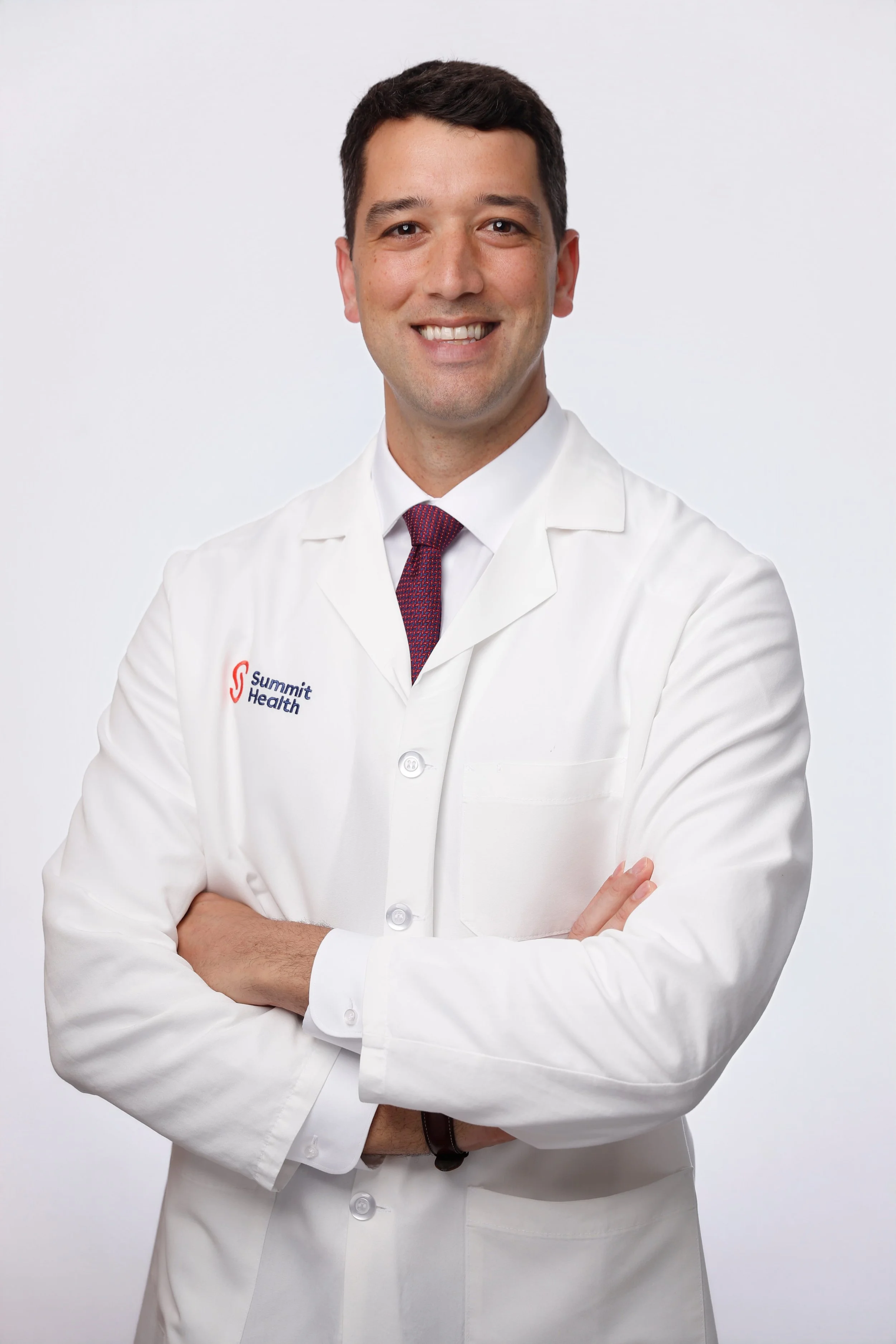 A male doctor wearing a white coat with Summit Health logo, smiling, standing against a plain white background with arms crossed.
