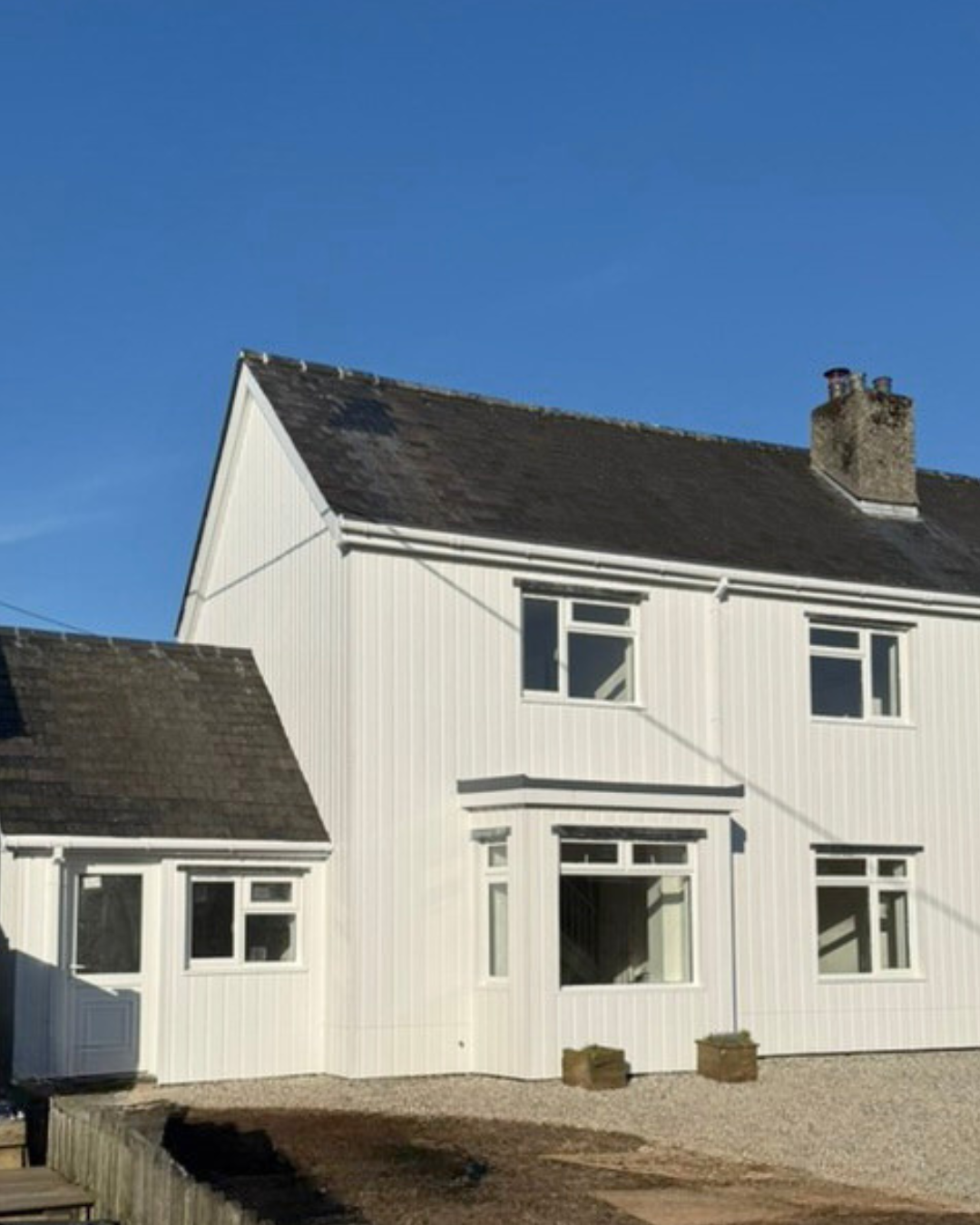 A two-story white cladded house with vertical siding, multiple windows, and a dark shingled roof against a clear blue sky.