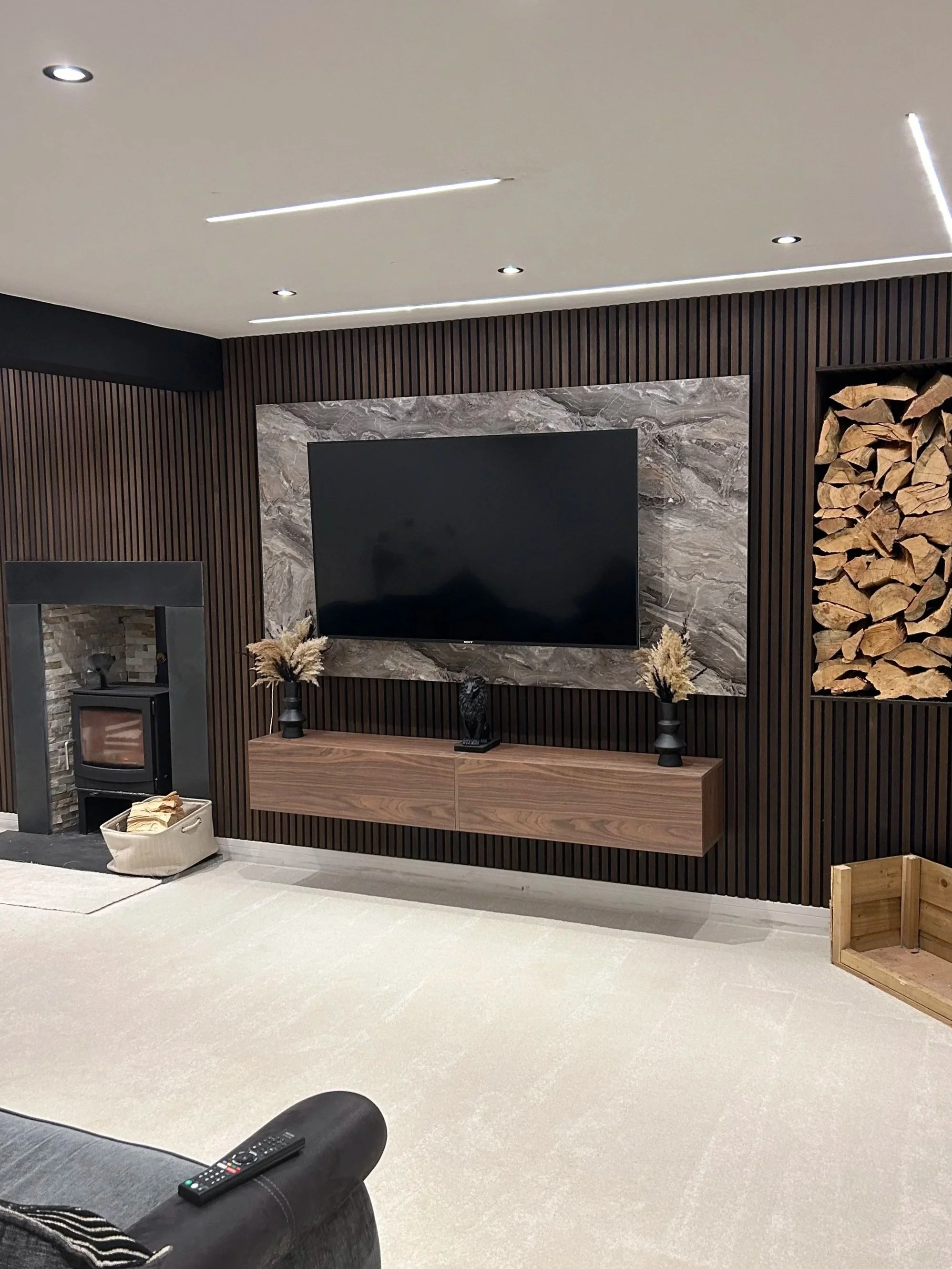 Living room with a mounted television on a marbled wall, wooden console beneath, and decorative vases with dried flowers. To the right, a wall with stacked logs for a fireplace or decor. Part of a black sofa and a remote control on it are visible in the foreground.