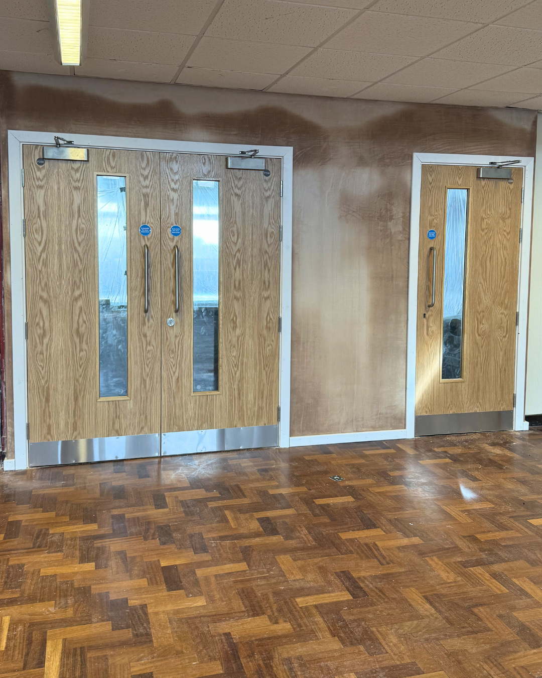 Two wooden doors with glass panels lead into a building under renovation, with a partially painted wall and wooden floor.