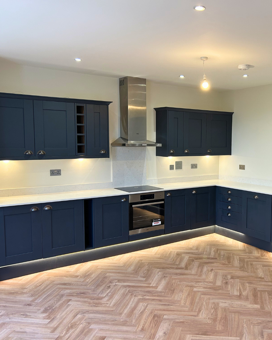 Modern kitchen with dark blue cabinets, white countertop, wooden herringbone floor, stainless steel oven and chimney, bright under-cabinet lighting, ceiling lights, and a bare bulb hanging from the ceiling. New kitchen fitted.