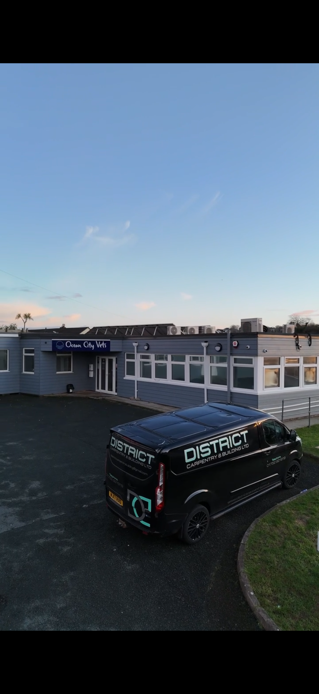 A veterinary clinic named Ocean City Vets with a dark blue sign and white lettering. A black work van with the word DISTRICT and the phrase Carpentry & Building LTD is parked in front. The building has grey siding, multiple windows, and a paved parking lot.