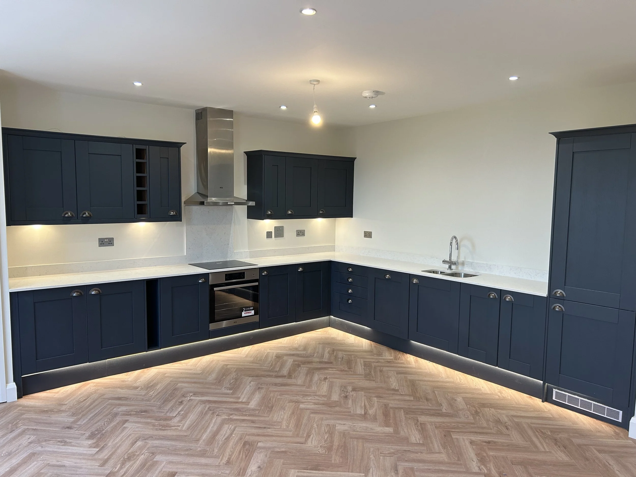 Modern kitchen with navy blue cabinets, stainless steel oven and exhaust hood, white countertops, and wooden herringbone flooring.