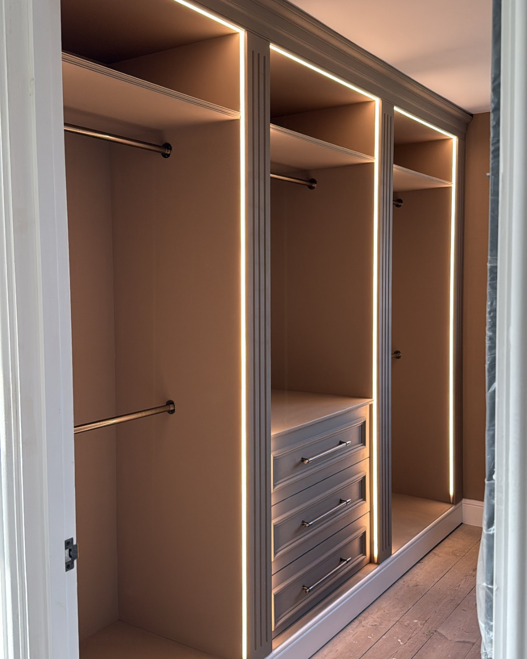 Modern built-in wardrobe with vertical LED lighting, multiple drawers, and hanging space, in beige and gray tones.