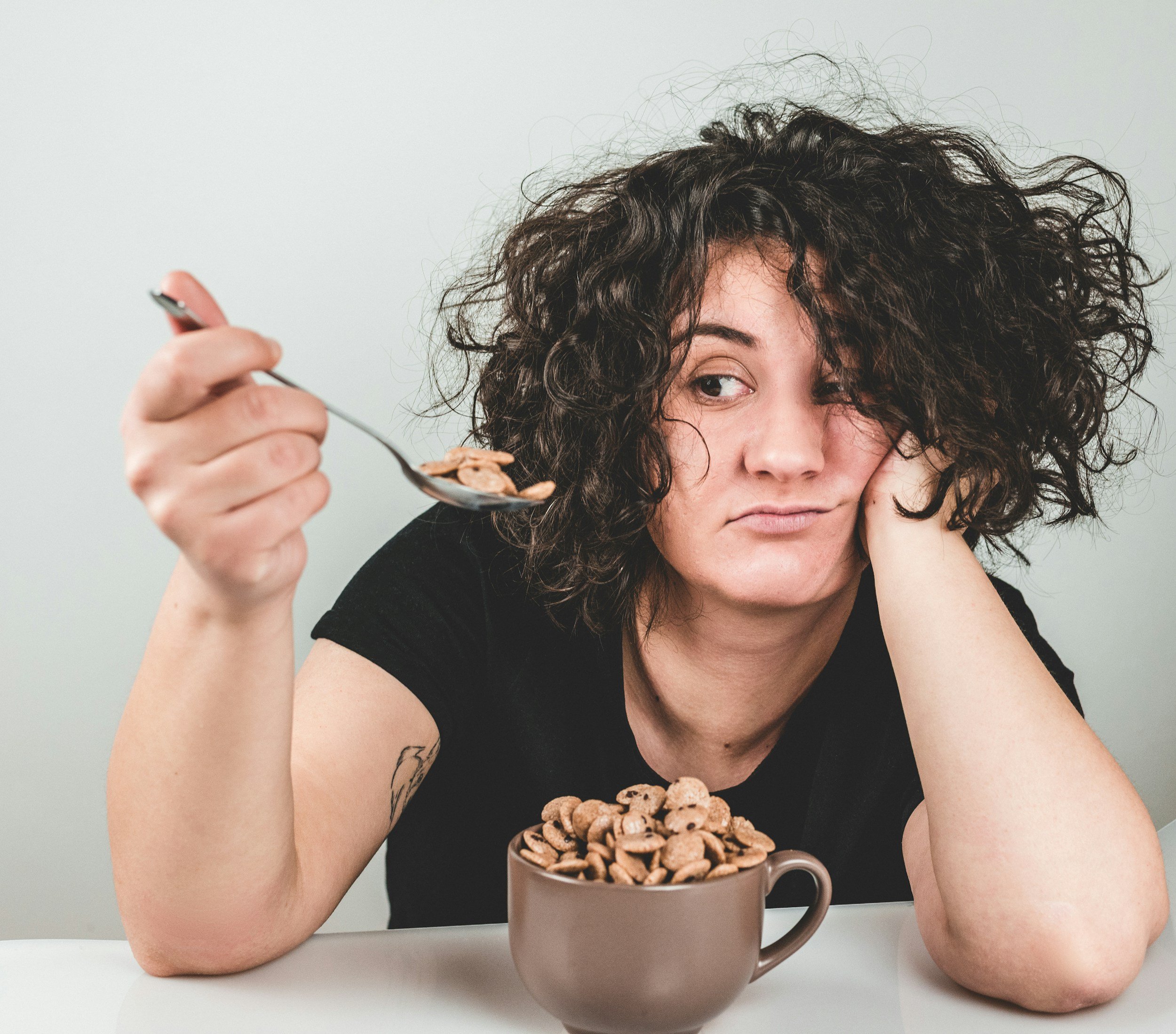Woman with curly hair holding a spoonful of cereal near her face, with a large mug of cereal and milk in front of her on a white surface.