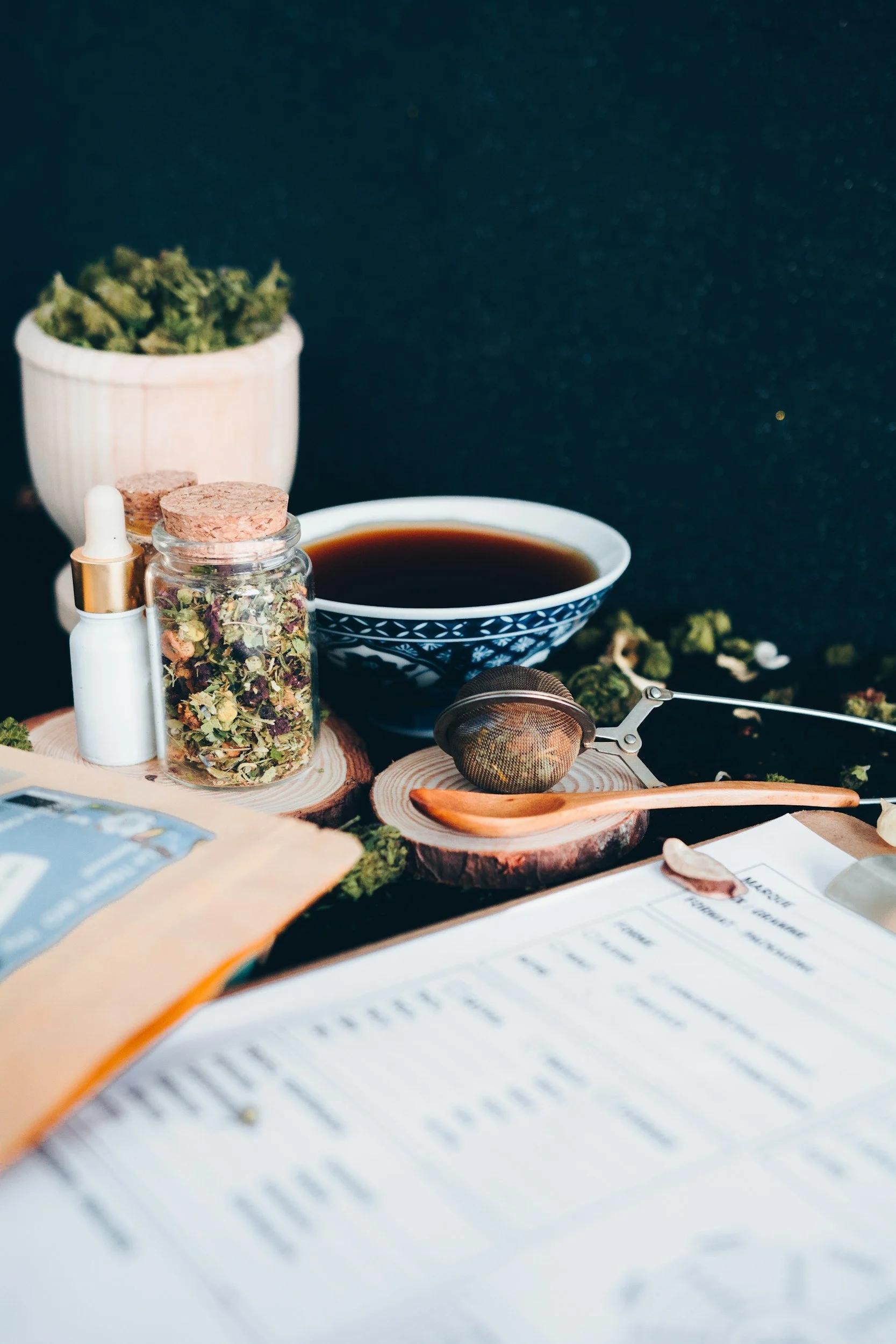 Variety of herbal treatment on a table, including dried cannabis buds, a jar of assorted dried herbs or flowers, a small dropper bottle, a bowl of tea or herbal infusion, a tea infuser, and some scattered dried herbs, with a dark textured background.