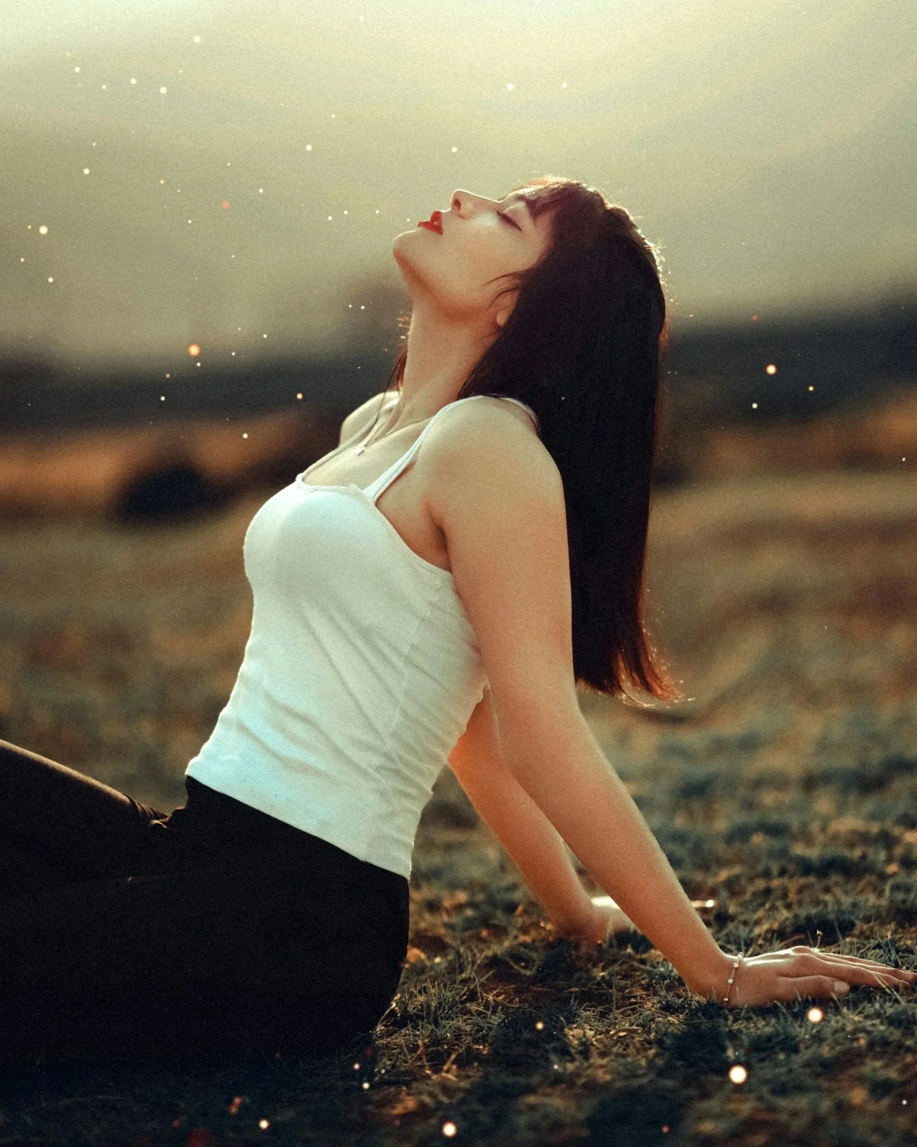 A woman with brown hair and red lipstick in a white tank top sitting on the ground outdoors, with one arm extended behind her supporting her and her head tilted back, during sunset with glowing particles in the background.