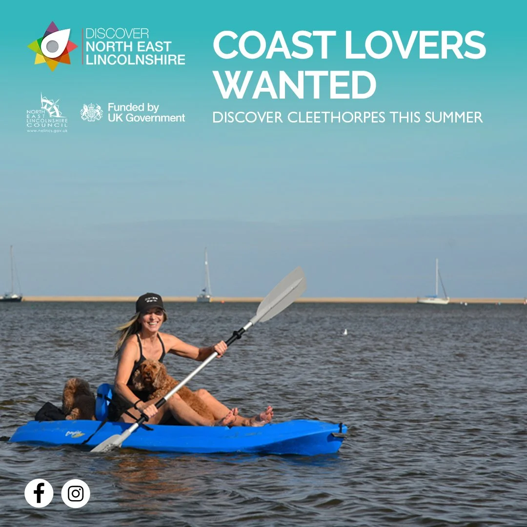 A woman kayaking on the water with a brown dog. The woman is smiling, holding a paddle, and wearing a black cap. The boat is a bright blue kayak. In the background are sailboats and a breakwater. The image is part of an advertisement seeking coast lovers to discover Cleethorpes this summer.