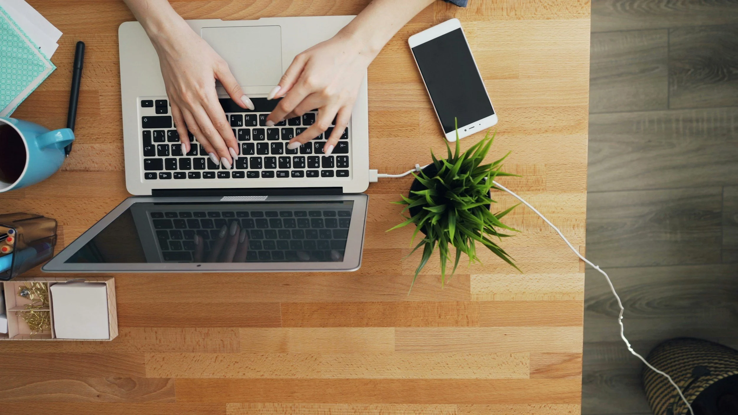 Top-down view of a wooden desk with a person typing on a silver laptop, a white smartphone with a black screen, a green potted plant, a blue mug with a beverage, a black pen, a small box with gold paper clips, a stack of teal cards, and a laptop computer.