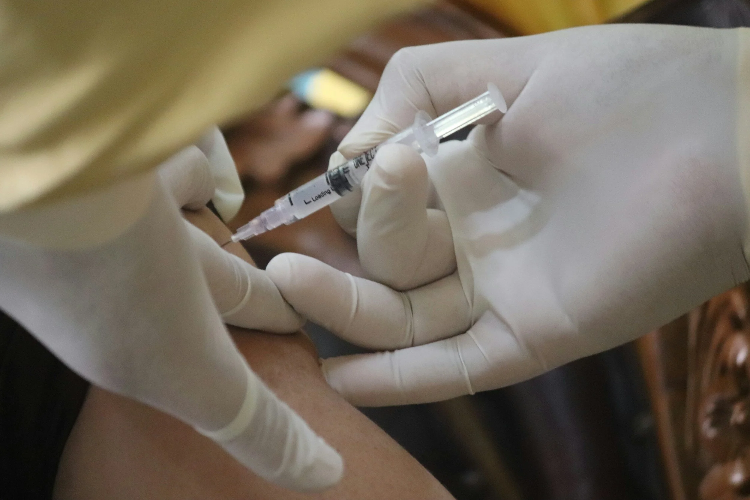 A person wearing white gloves administering a shot with a syringe into someone's arm.