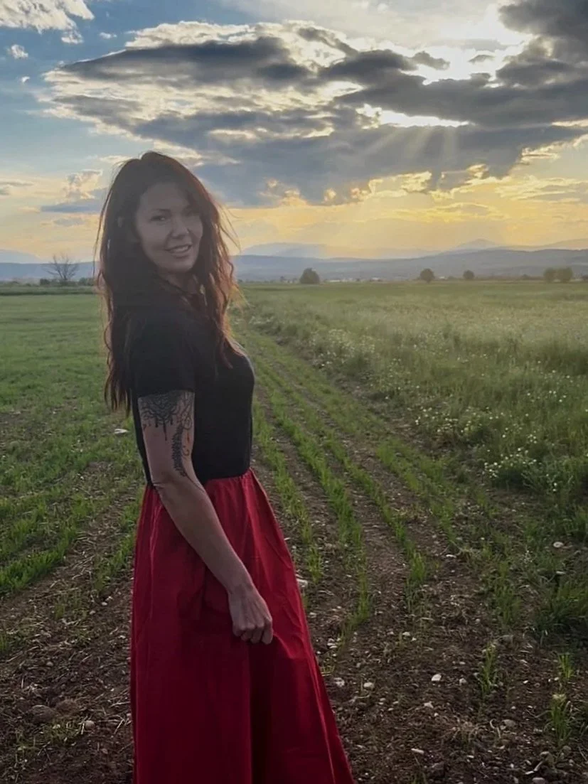 A woman standing in a field at sunset, with a cloudy sky overhead. She has long hair, wears a black shirt and a red skirt, and has a tattoo on her left arm. She is smiling and looking at the camera.